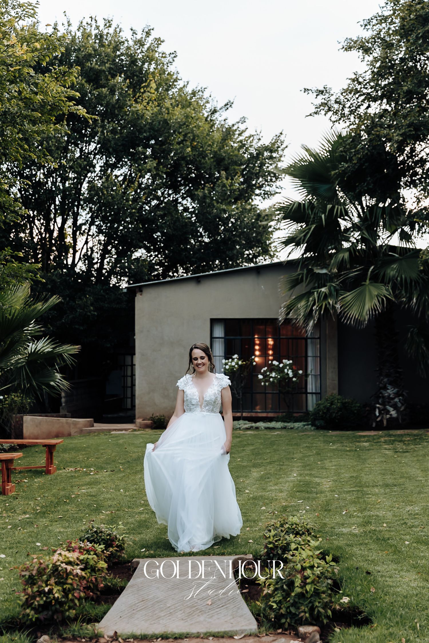 Bride in a flowing white lace wedding gown walking across a green garden lawn at an outdoor venue.