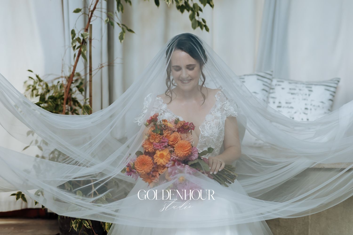 Bride in lace wedding dress holds vibrant orange bouquet beneath flowing veil indoors.
