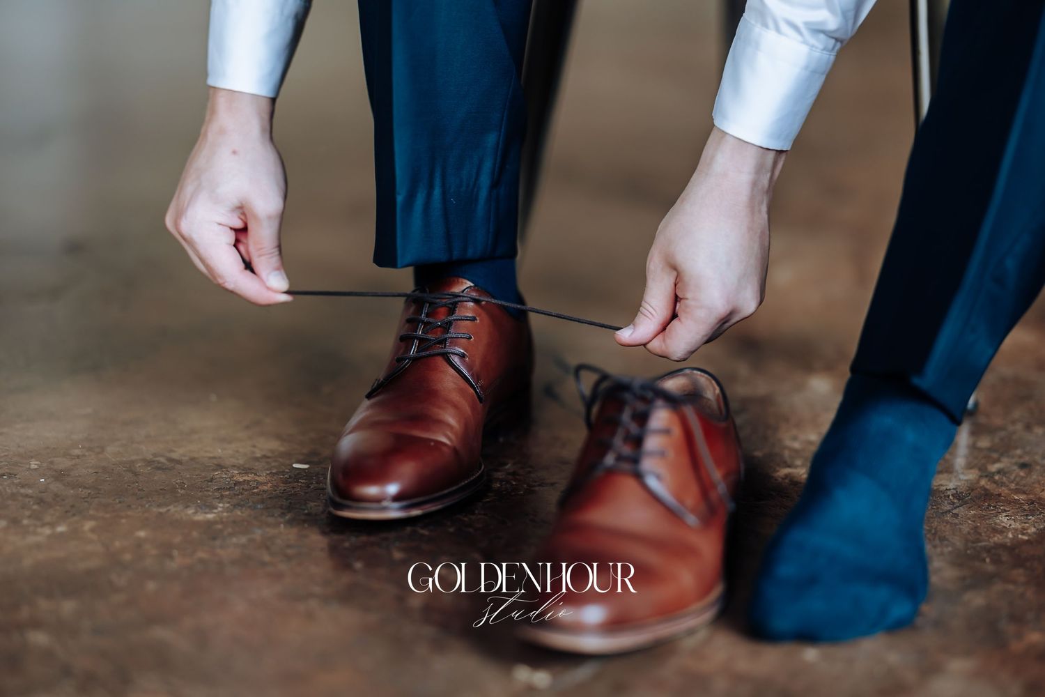 Groom tying brown leather dress shoes with blue socks on a concrete floor before the wedding ceremony.