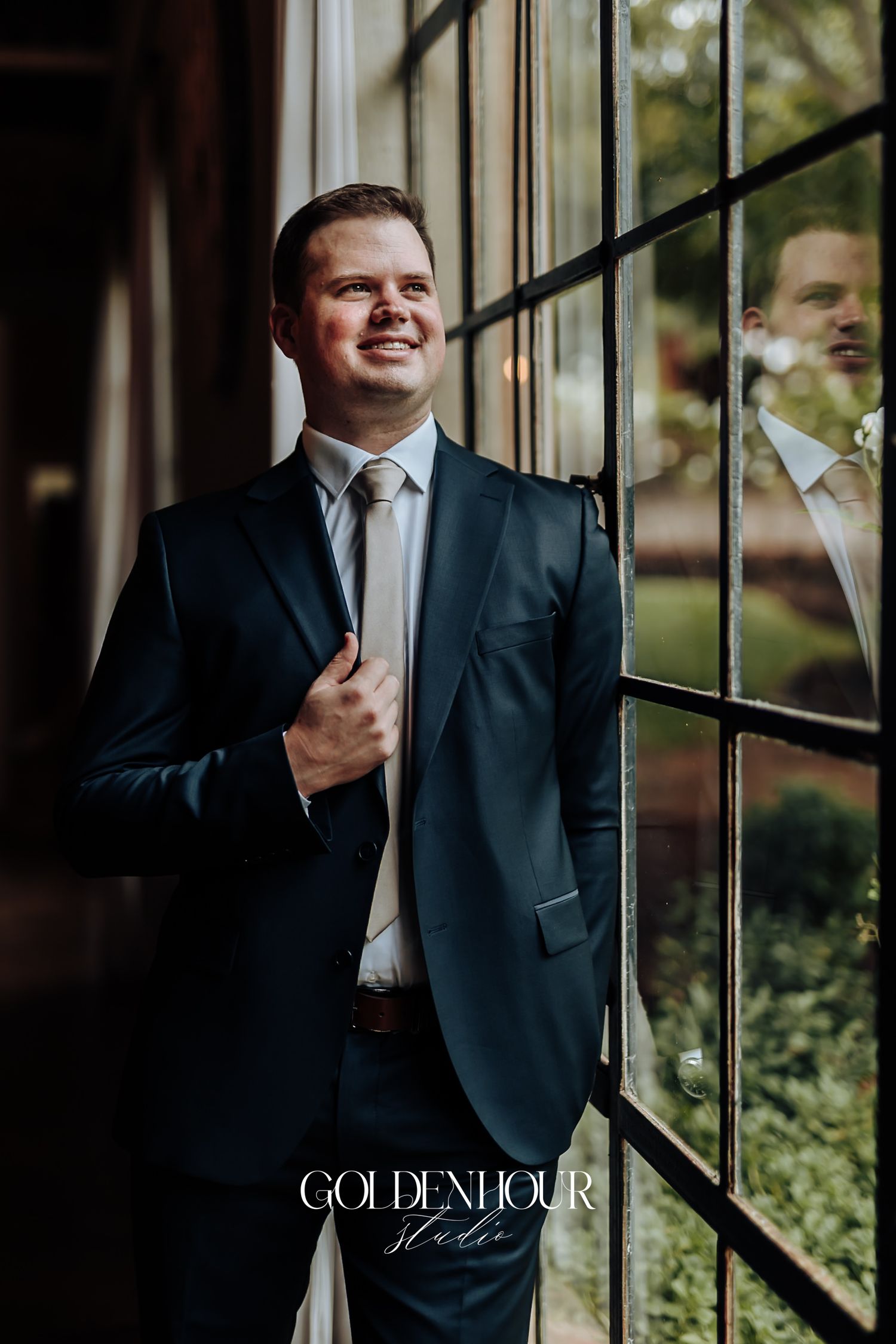 Smiling groom in navy suit and silver tie posing by large windows with natural light reflection.