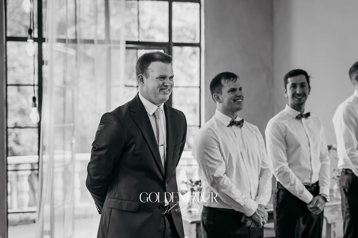 Black and white photo of groom smiling with groomsmen in bow ties at wedding ceremony.