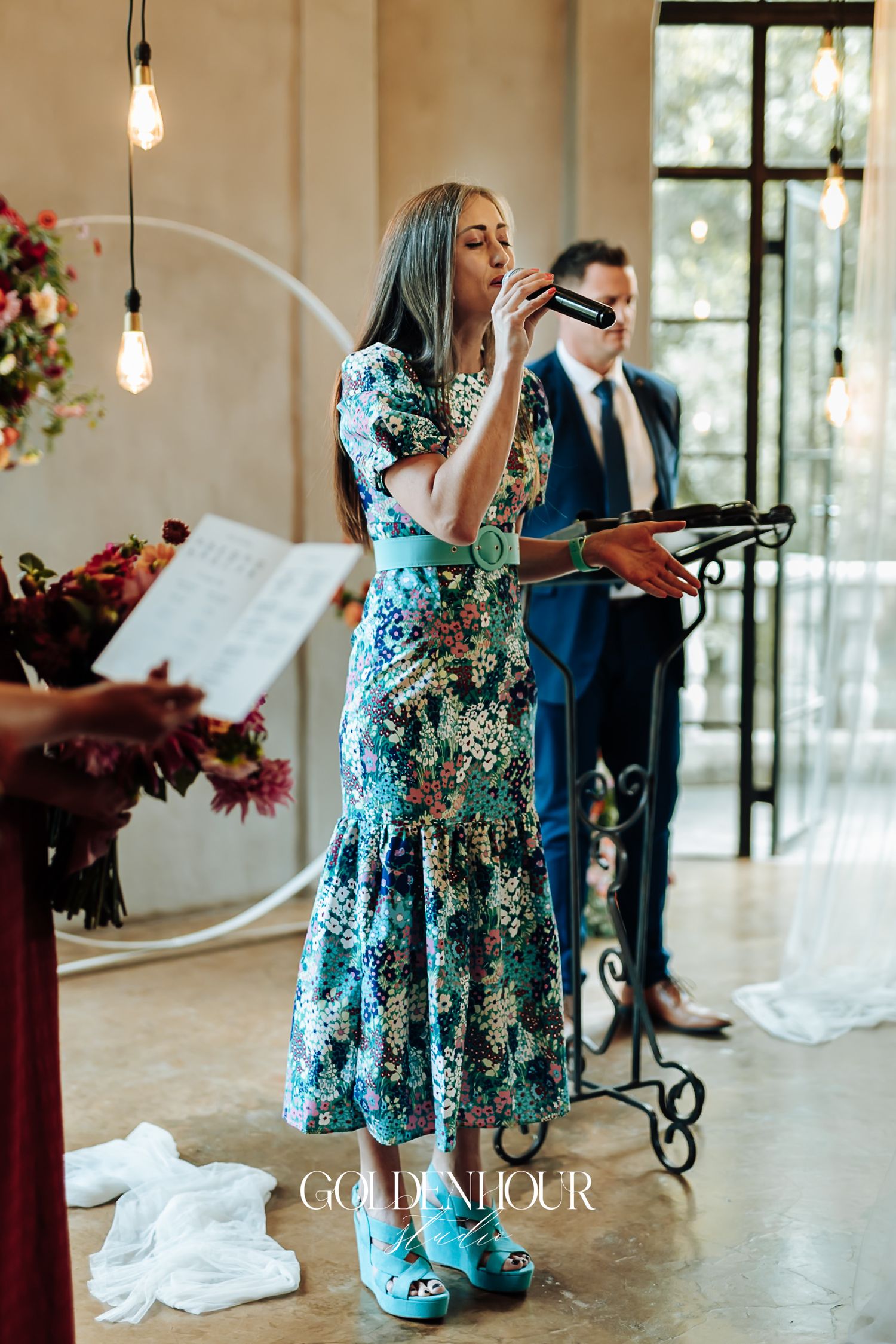 Wedding officiant in colorful floral dress speaking into microphone during indoor ceremony.