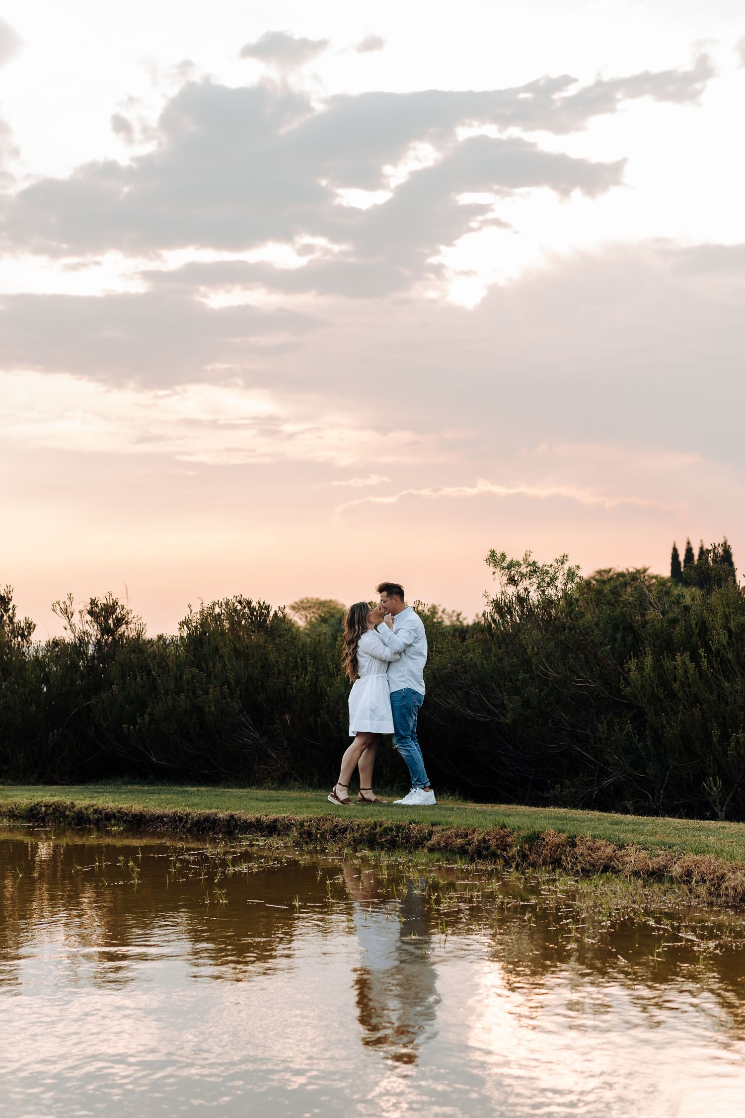 Couple embracing romantically by a reflective pond at sunset, surrounded by lush green trees under a pastel sky.