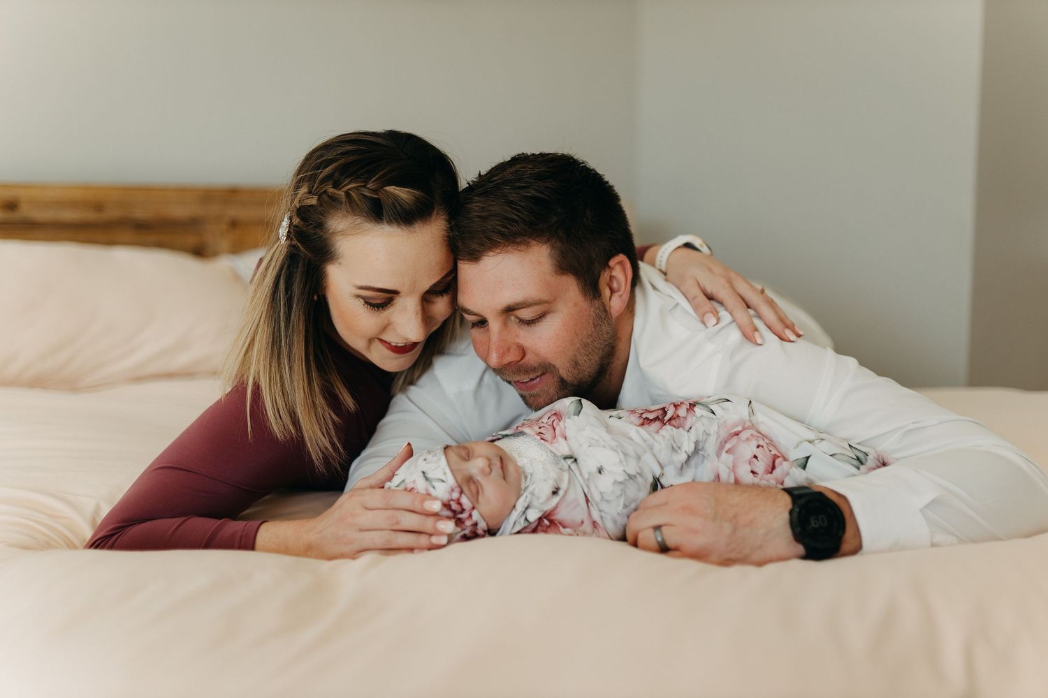 Parents lying on bed gazing lovingly at their swaddled newborn baby between them.