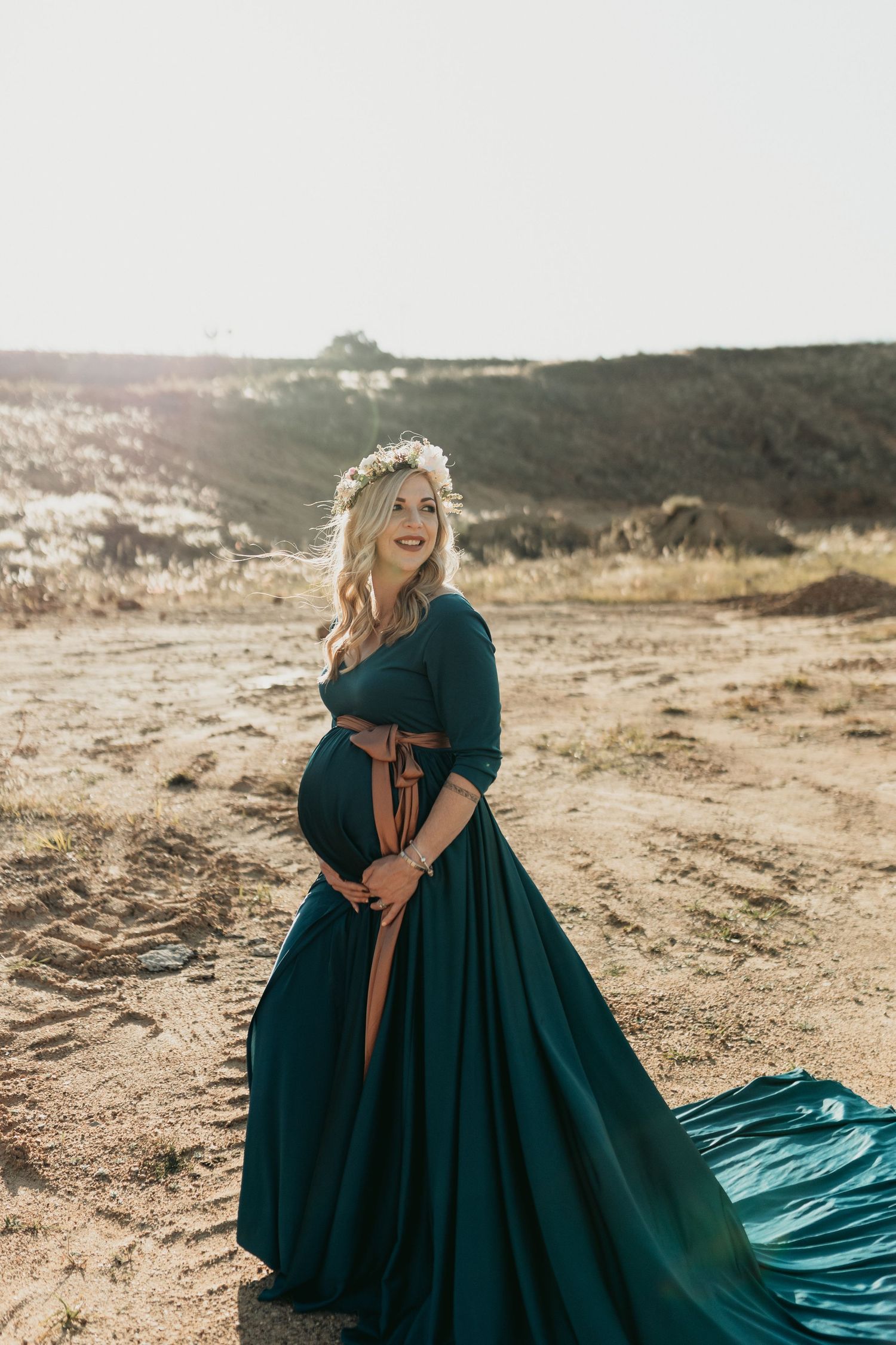 Expectant woman in flowing teal dress and floral crown poses against mountain landscape at sunset.