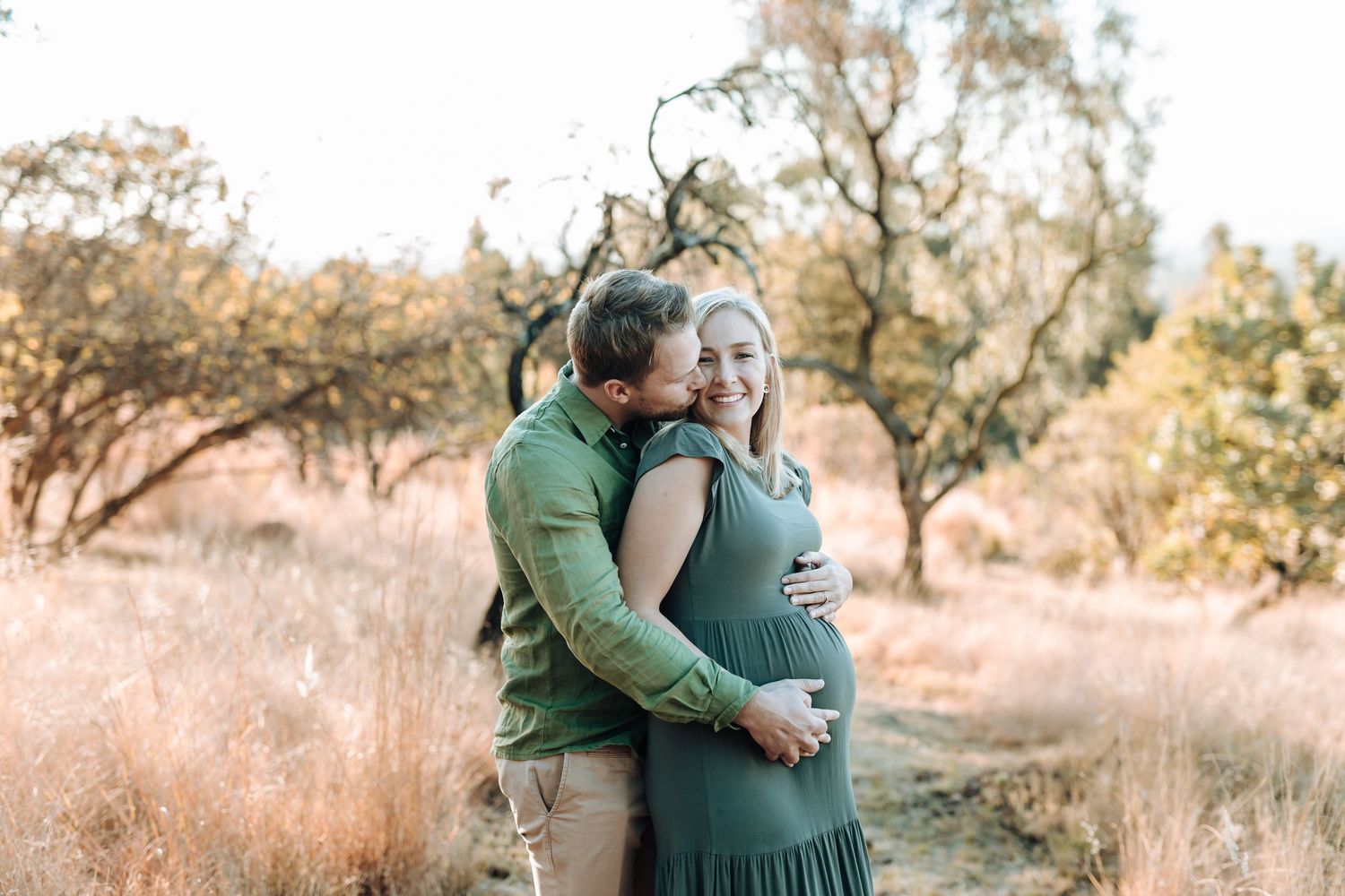 A romantic couple in formal green attire embraces outdoors in a sun-dappled wooded setting during golden hour.