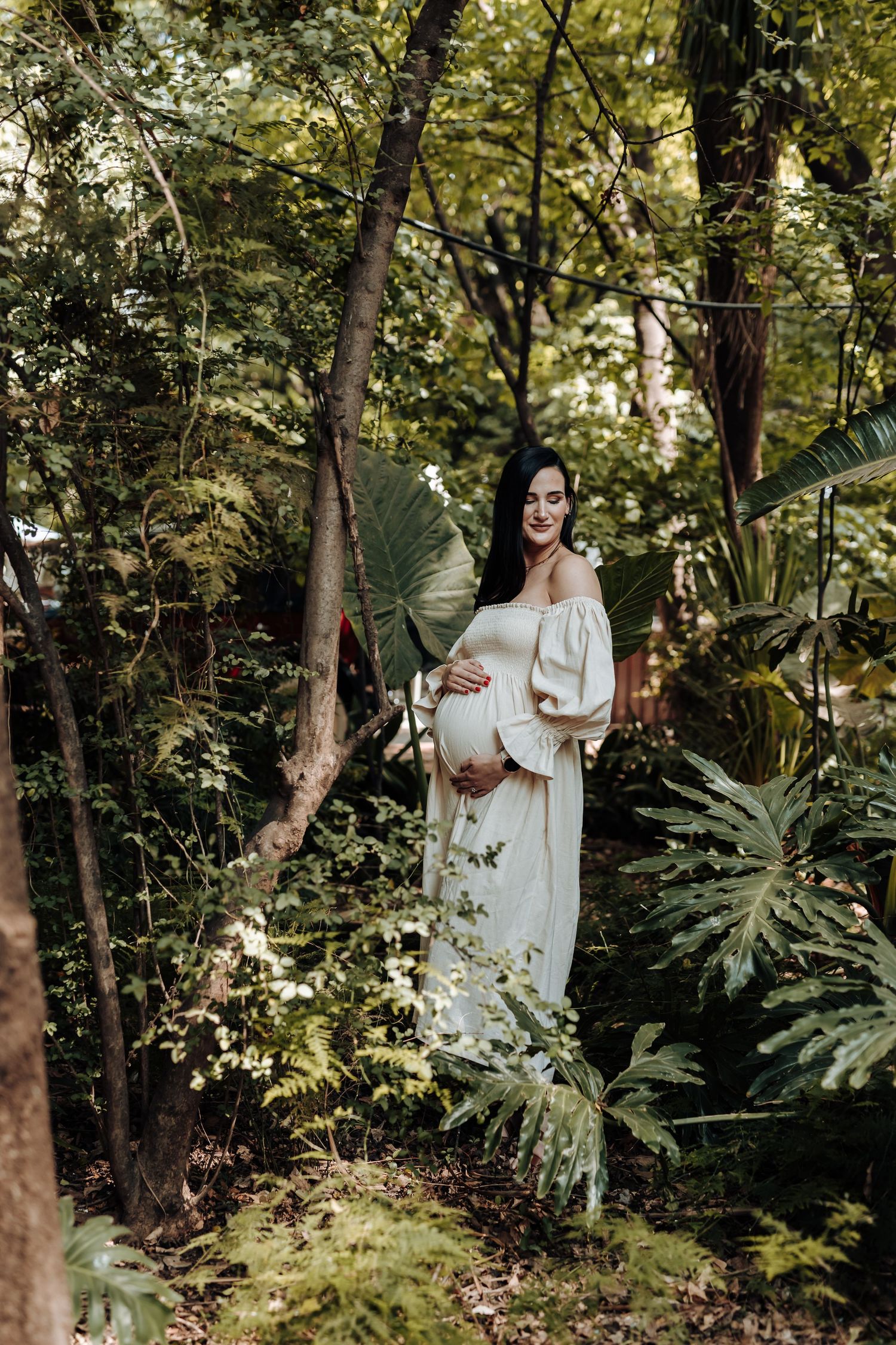 Expectant couple poses for maternity photos in a sunlit forest setting wearing white clothing.