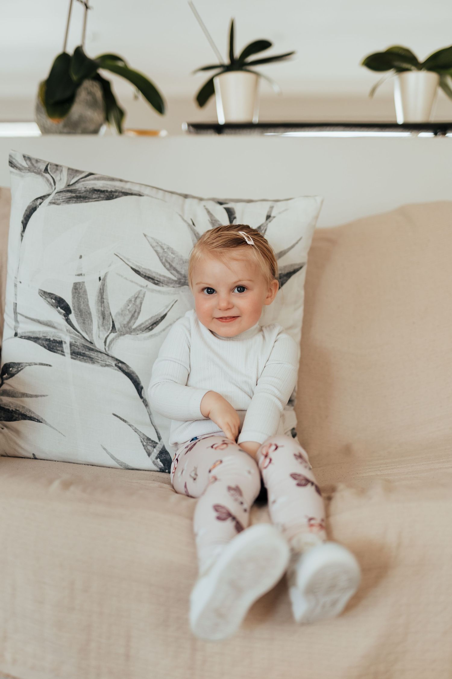 Baby relaxing on beige couch with floral decorative pillow in natural light setting.