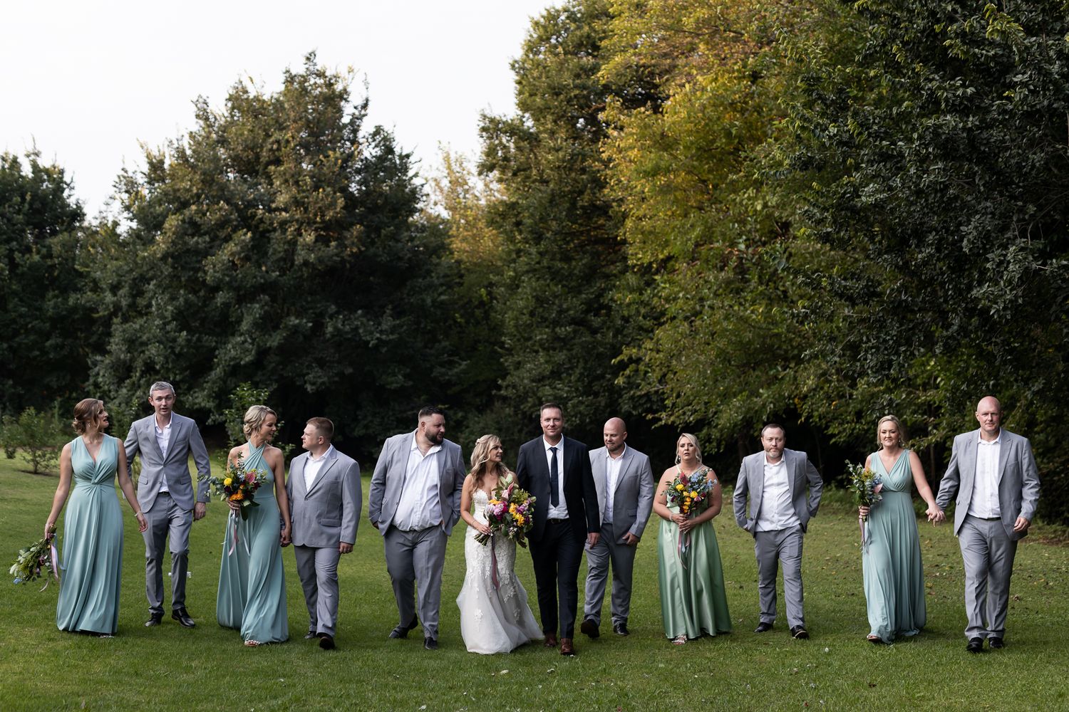 Wedding party in formal attire poses on grass with trees in background wearing light blue and gray colors.