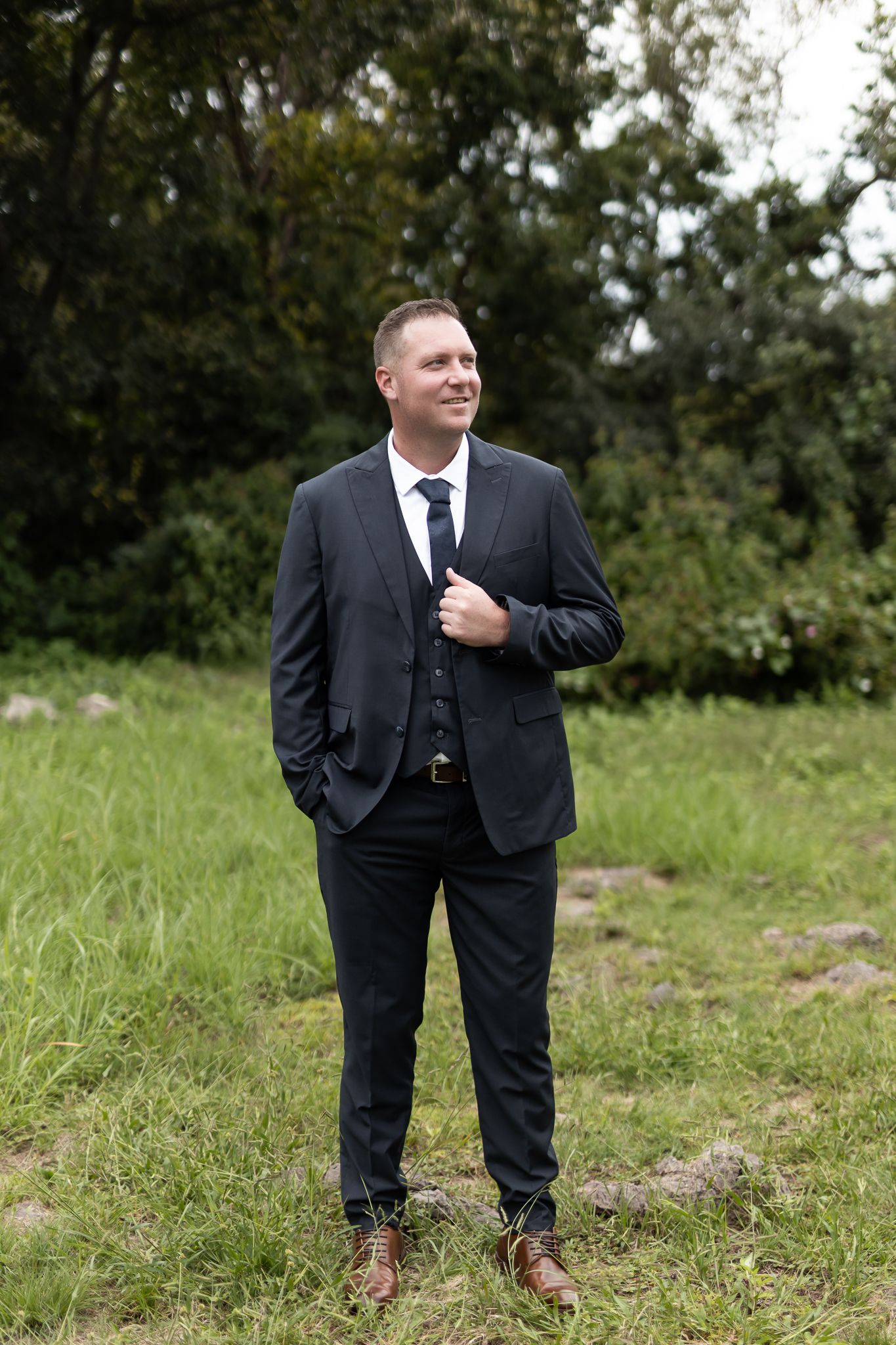 Groom in dark suit smiles while standing in grassy field with trees in background.