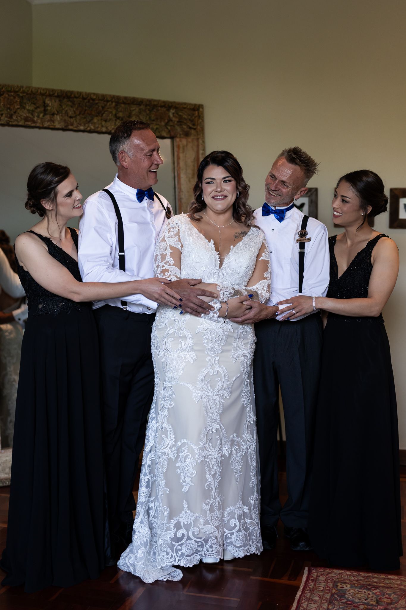 Wedding party in black attire gathers around bride in lace gown for pre-ceremony photos.