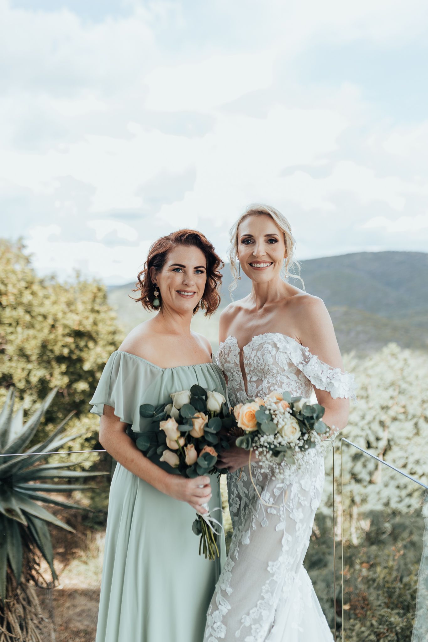 Bride in off-shoulder lace gown poses with bridesmaid in sage dress against mountain backdrop.