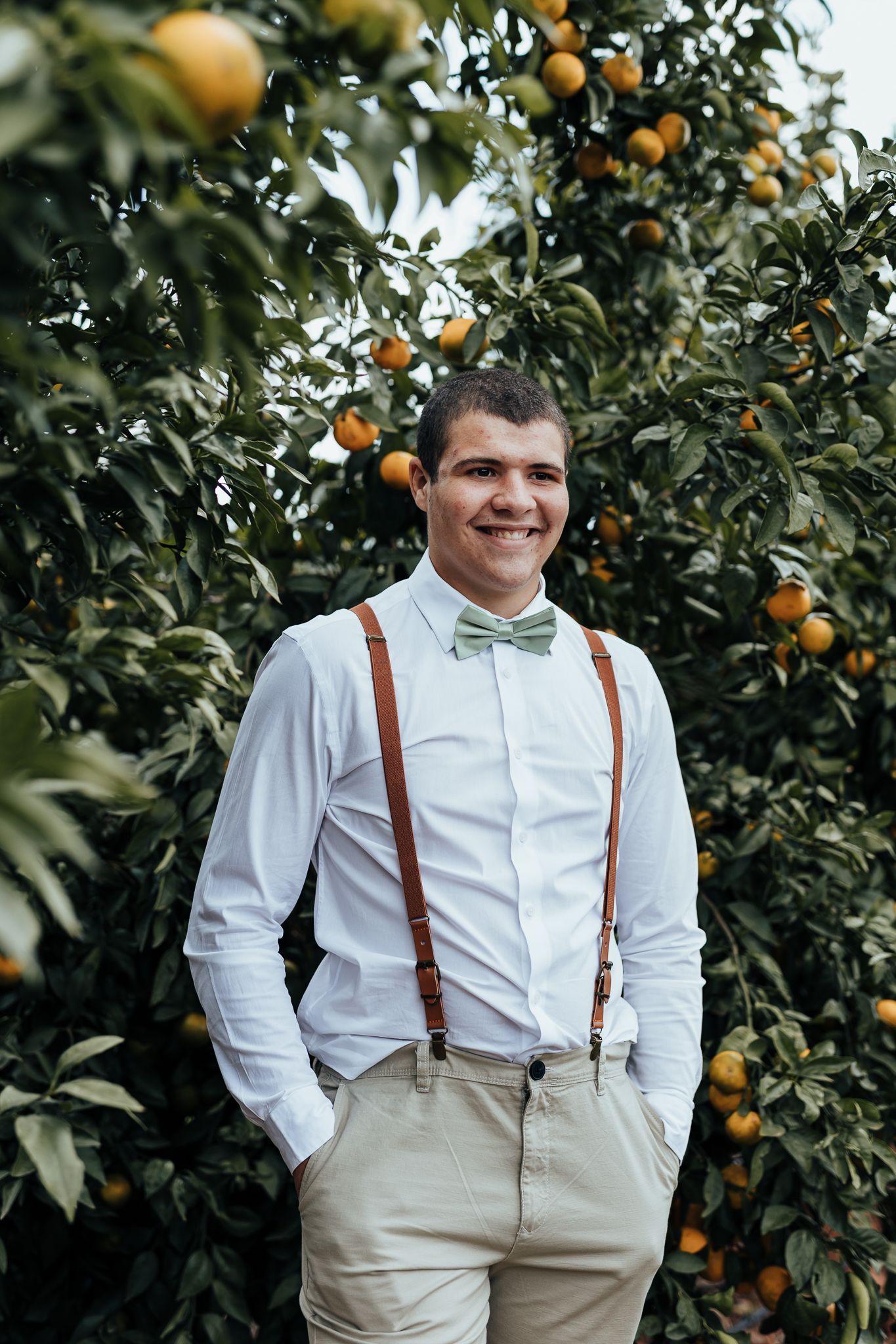 Wedding attendee wearing suspenders and bow tie smiles in front of orange trees.