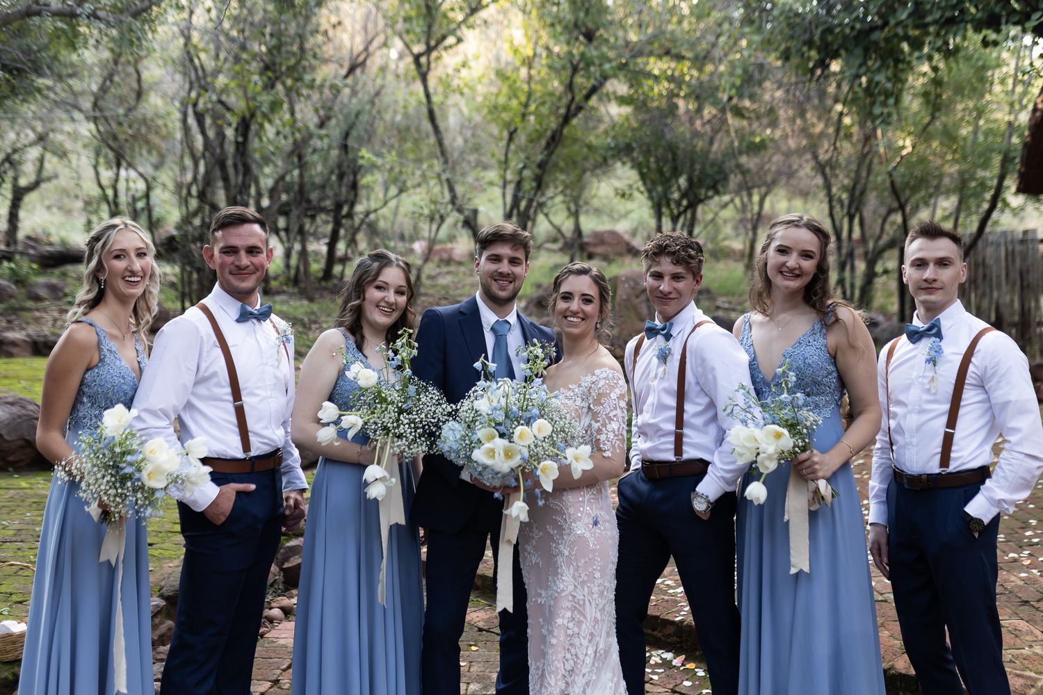 Wedding party members in blue dresses and suspenders pose together for formal group photo outdoors.