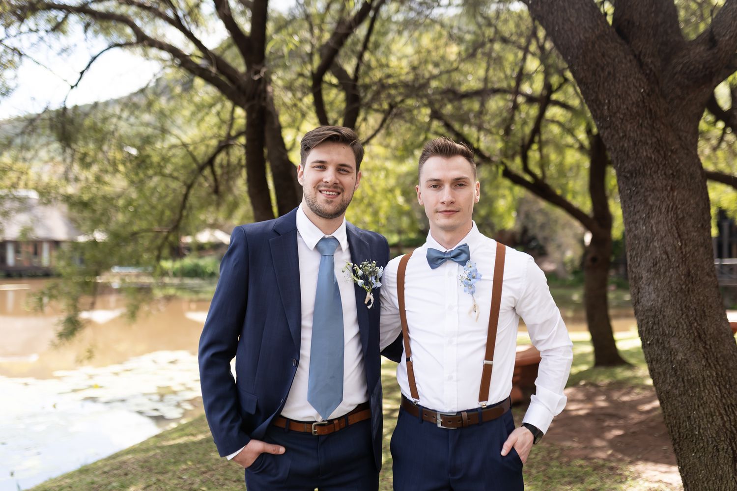 Two men in coordinated wedding attire smiling by waterfront.