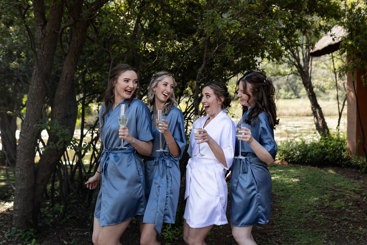 Group in matching robes shares laughs while holding champagne glasses in a garden.