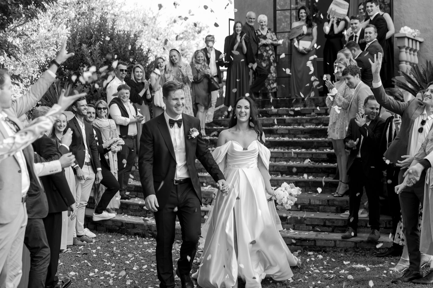 Newlyweds descending stone steps while guests shower them with flower petals in black and white.