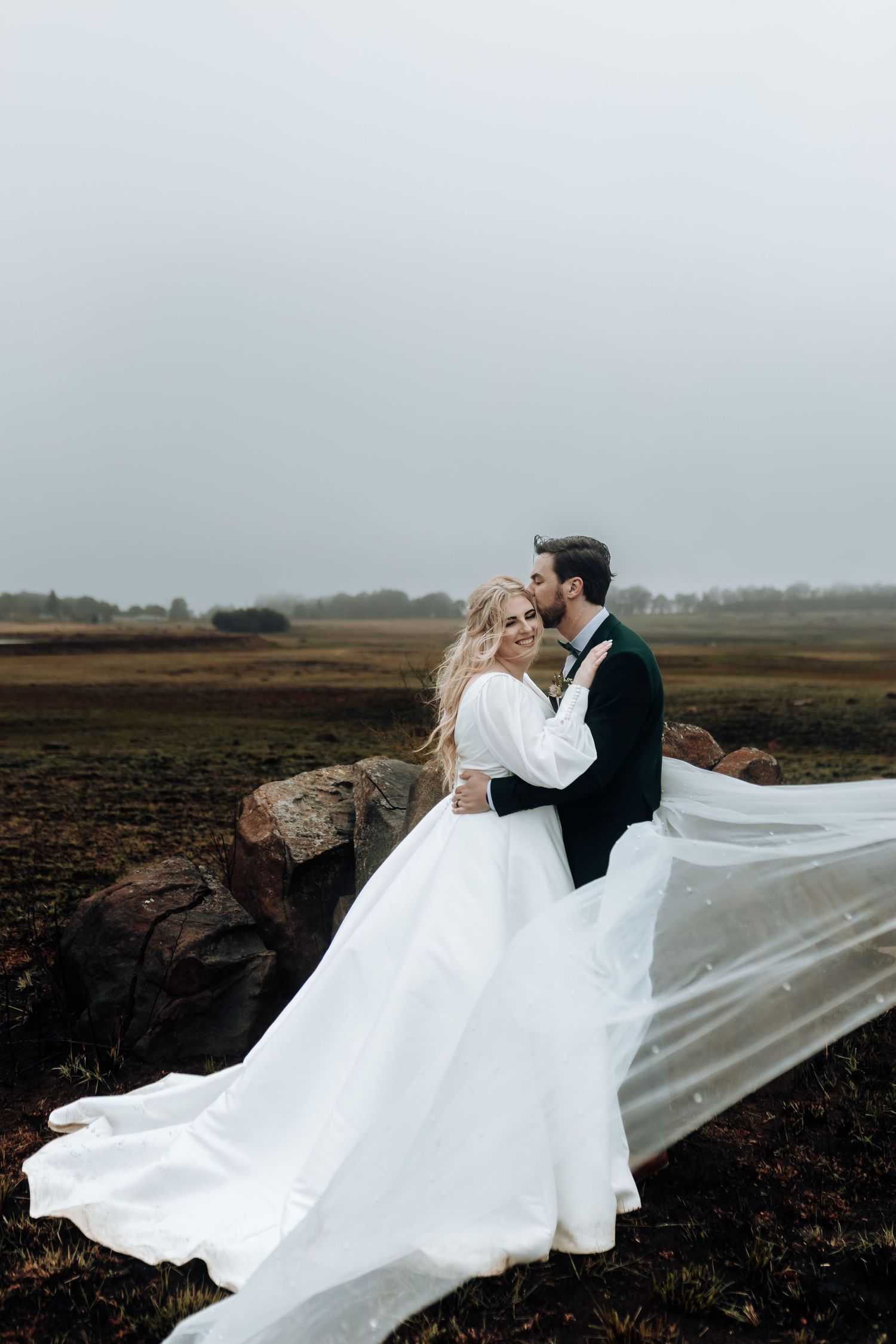 Dramatic wedding portrait with flowing veil against moody sky and rustic landscape.