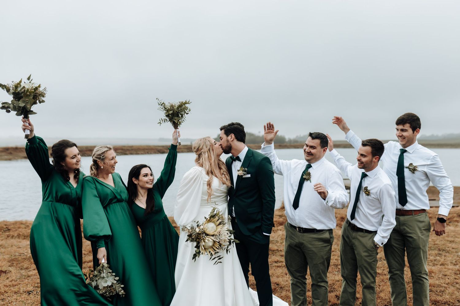 Wedding party celebrates enthusiastically by water with bridesmaids in emerald dresses.