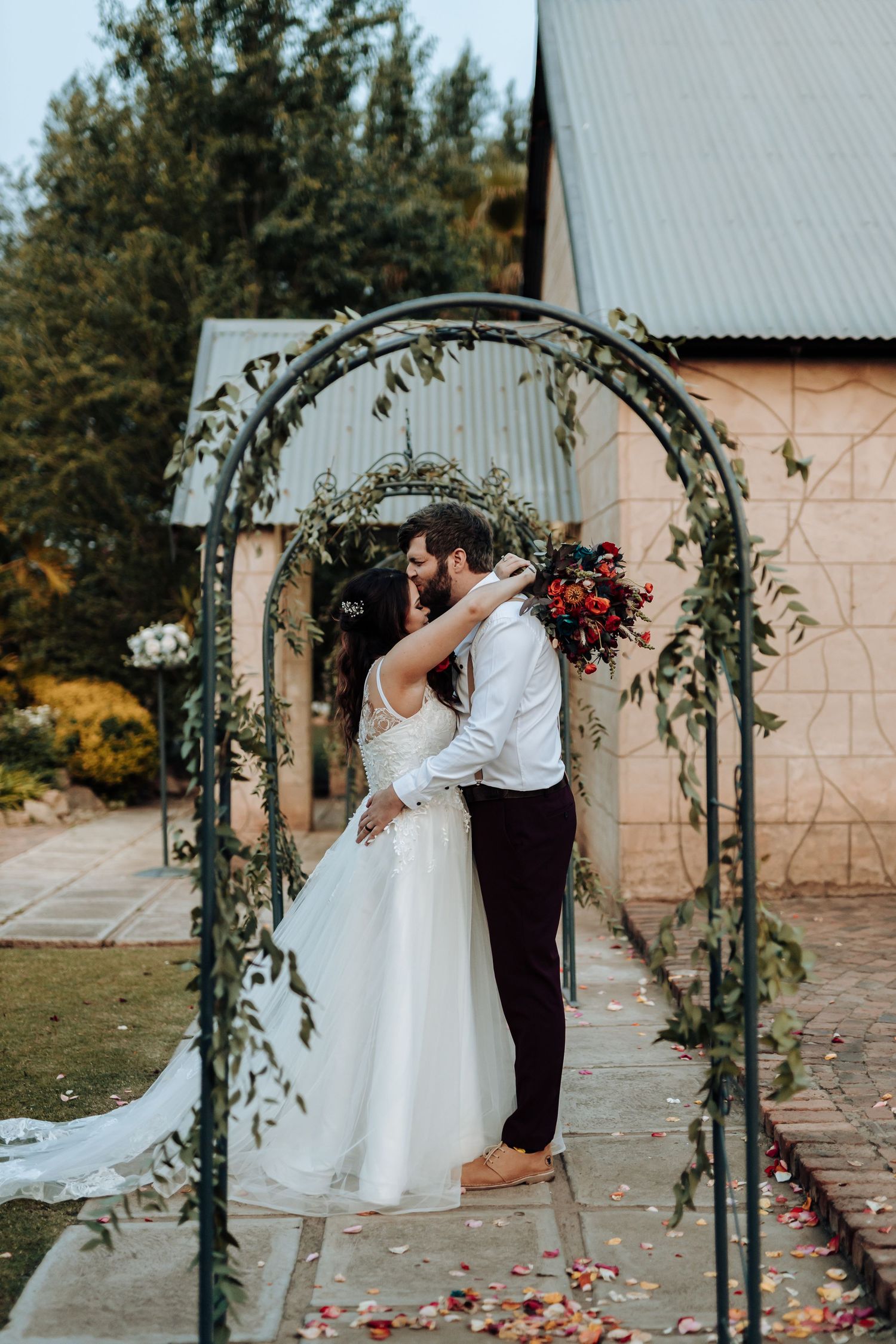 A wedding moment under an ivy-covered metal arch with pink stone building in background.