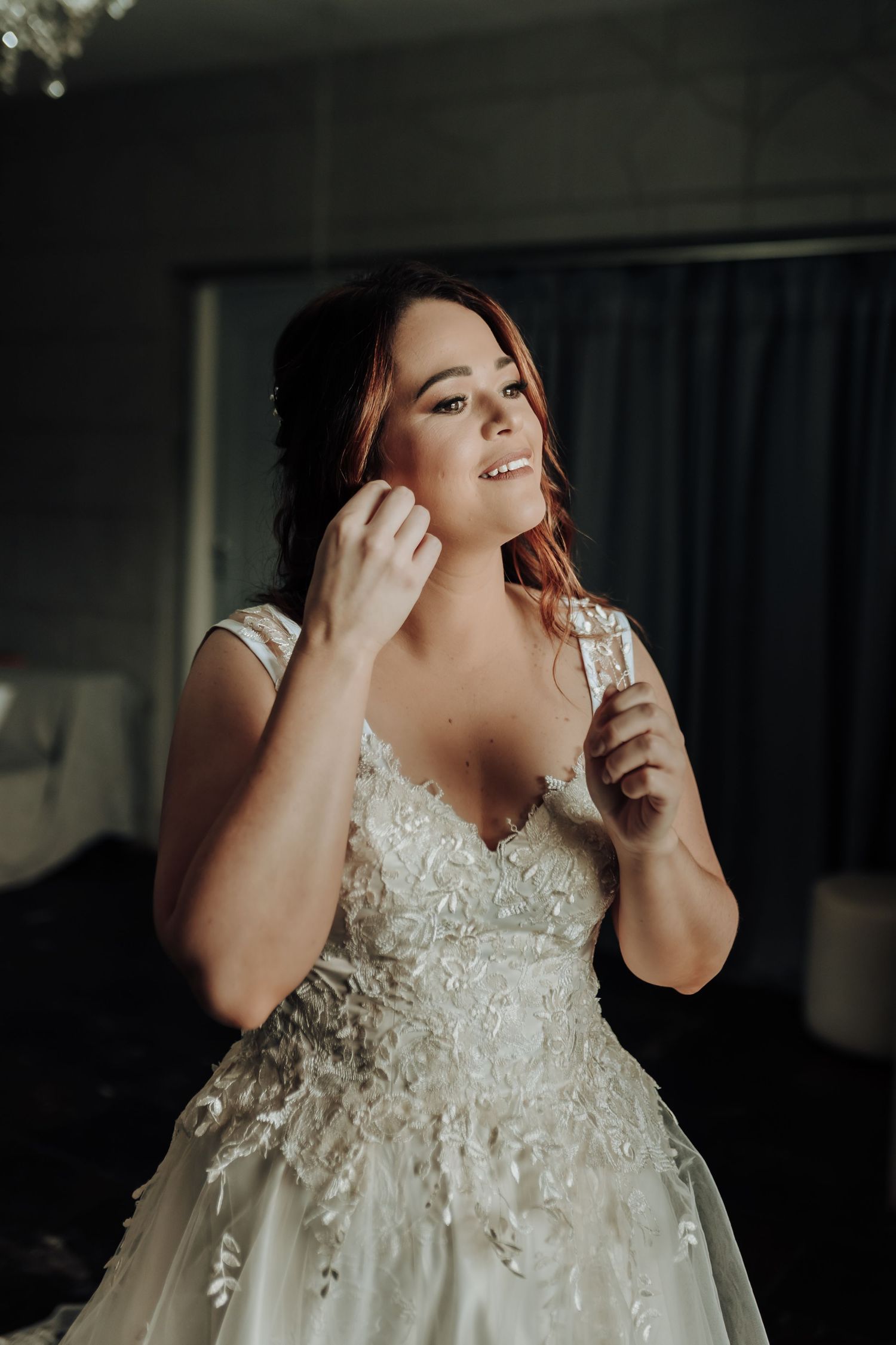 Bride in an embellished white wedding gown puts on earrings while getting ready.