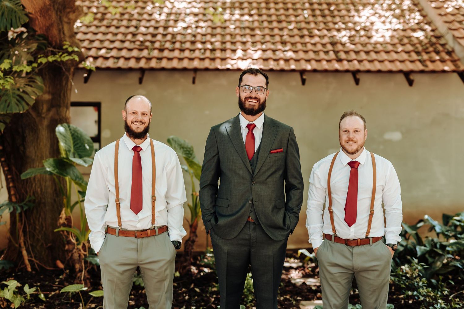 Three men in wedding attire stand together, with matching red ties and contrasting gray suit and white shirts.