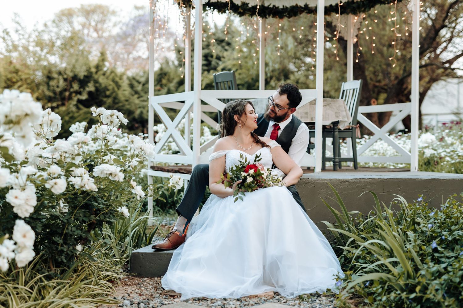 Wedding couple embracing on a bench in a white gazebo surrounded by lush gardens.