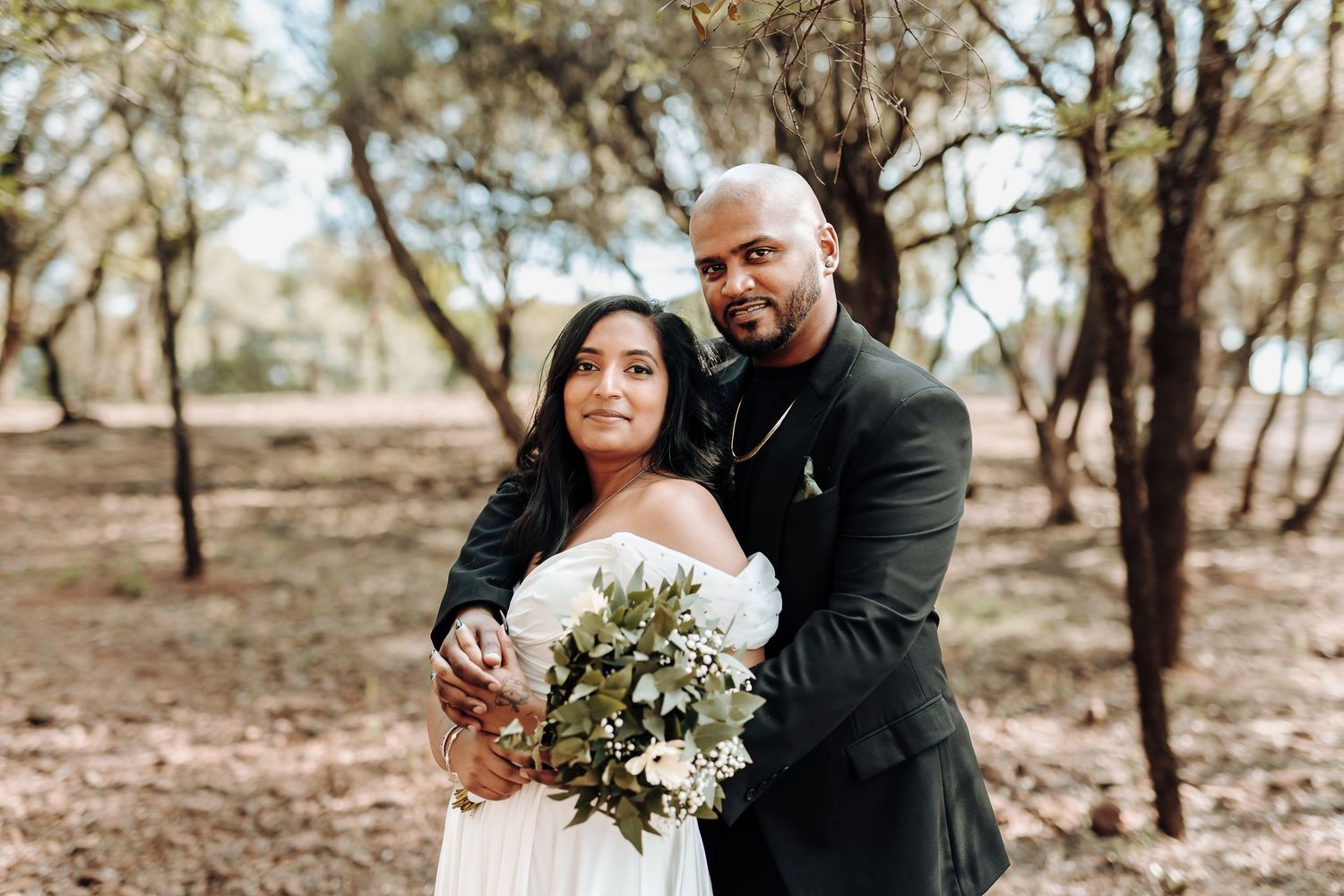 A couple in wedding attire embrace while holding a floral bouquet in a rustic outdoor setting.