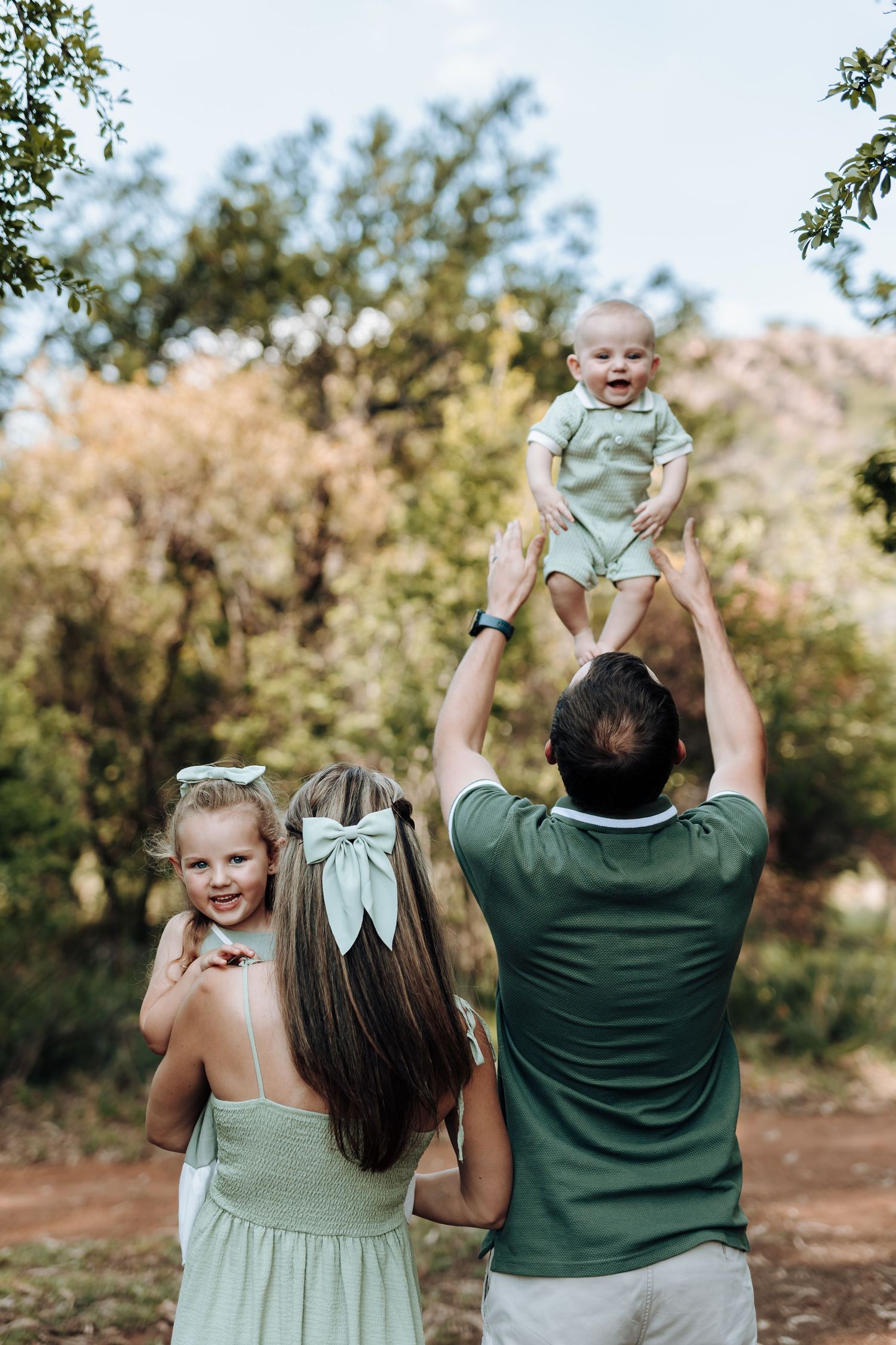 A candid family moment showing people in sage green outfits playing with an infant against a natural outdoor backdrop.