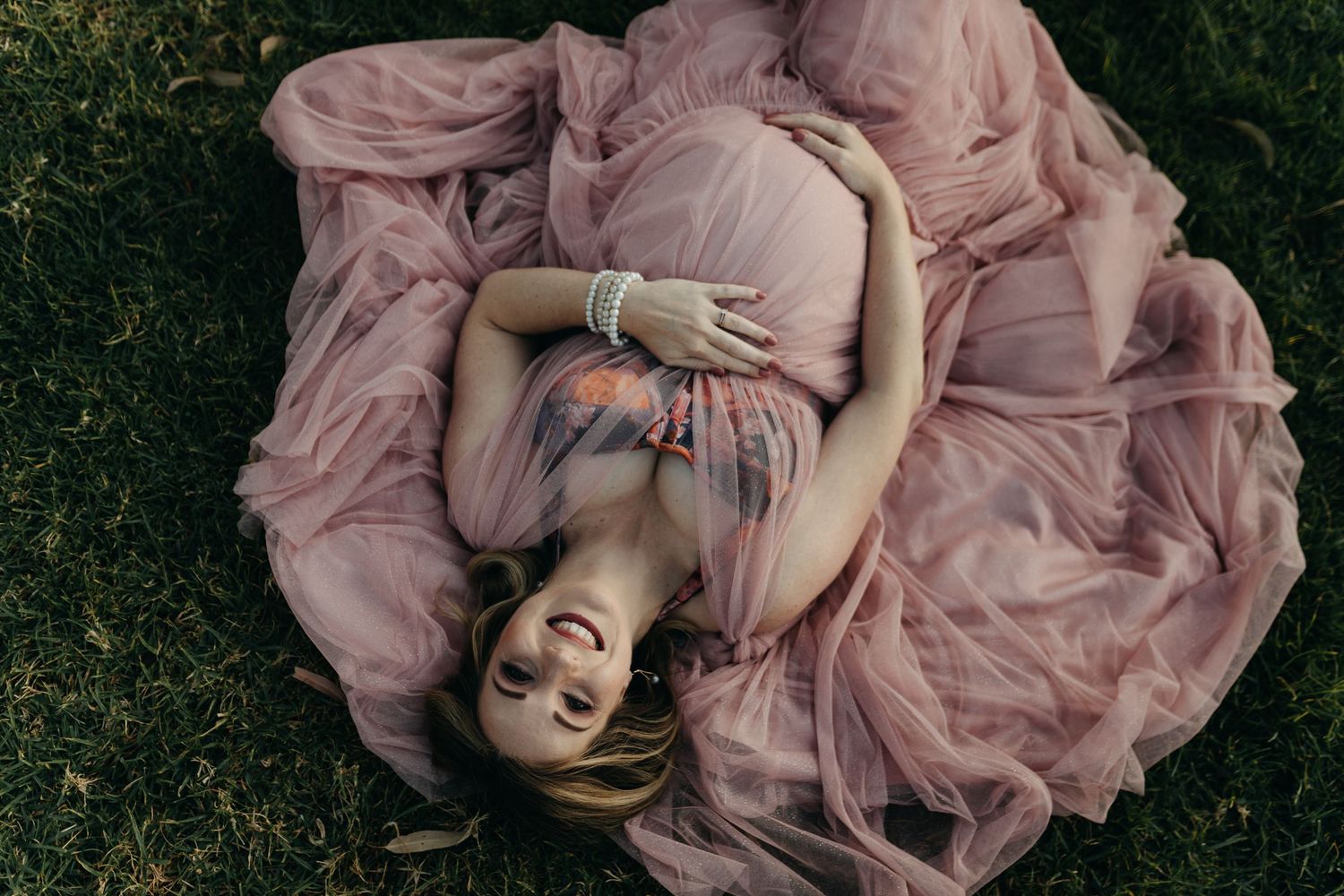 A person in a flowing dusty pink gown lies on grass, creating a circular shape with the fabric spread out around them.