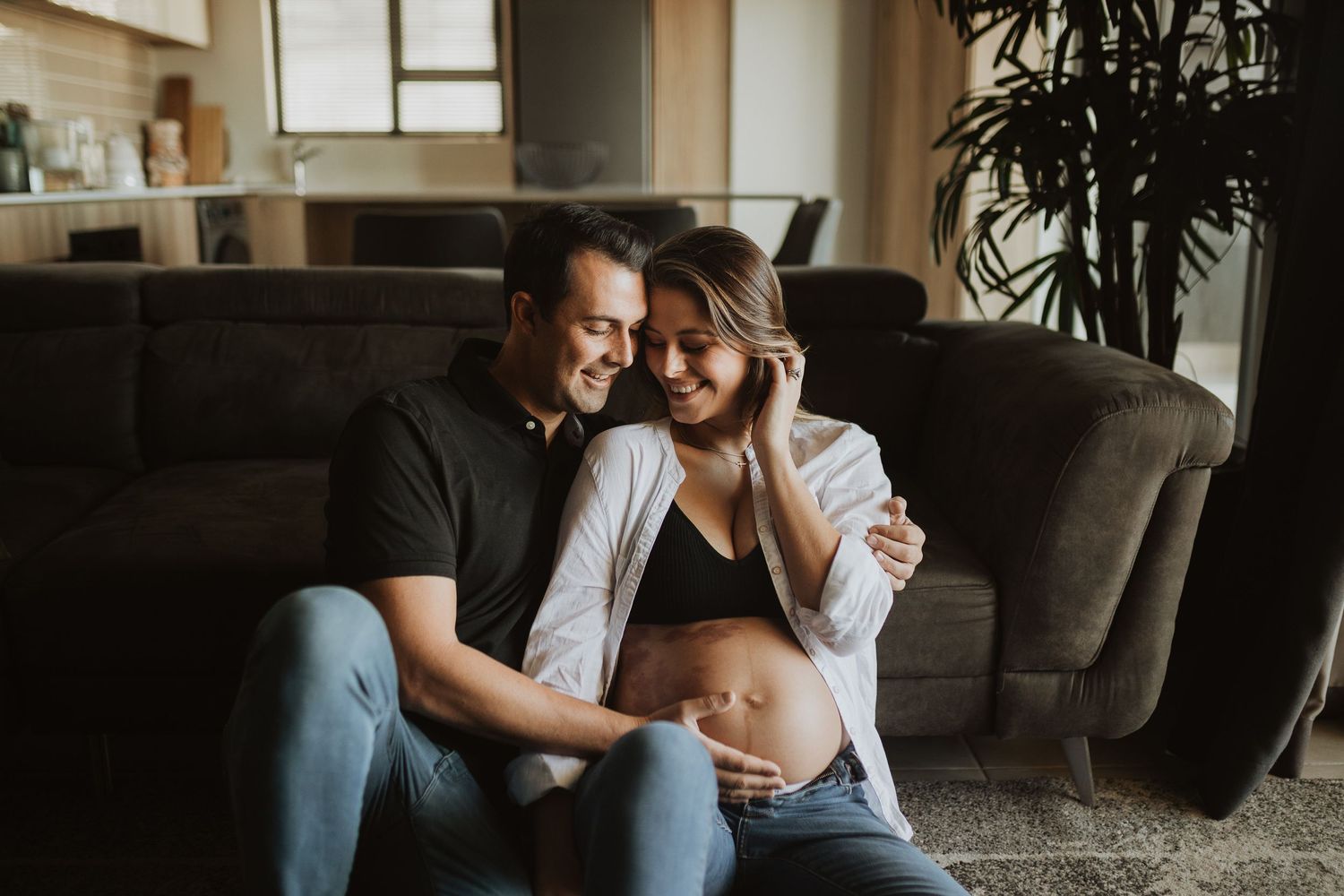 Couple embraces intimately on grey couch during indoor lifestyle photoshoot.