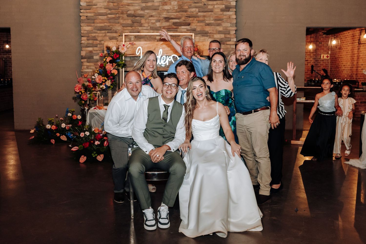 Wedding party poses for a fun group photo in a reception venue with brick accent wall.