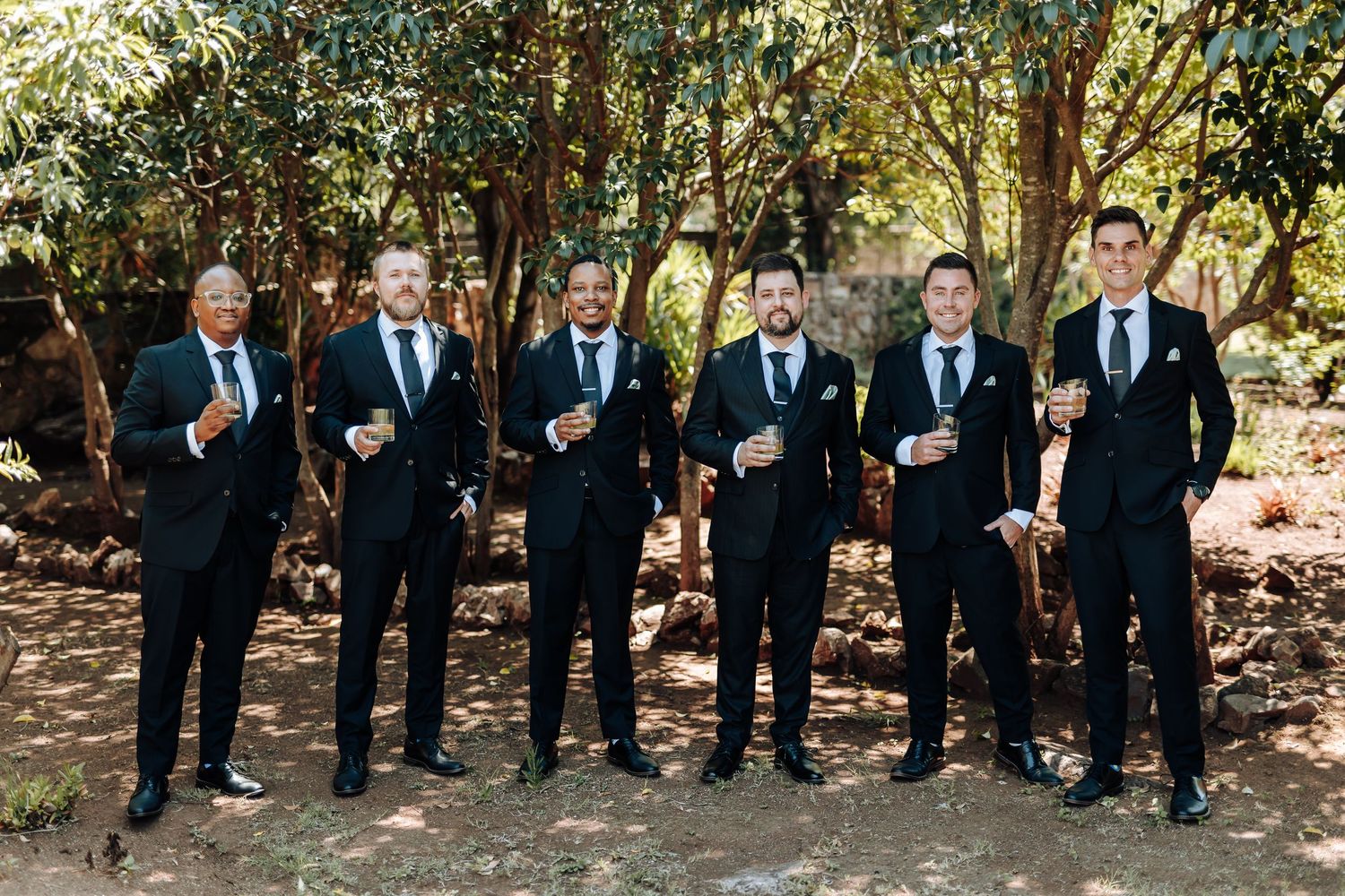 Groomsmen in dark suits pose together in a line outdoors with trees and natural lighting in the background.