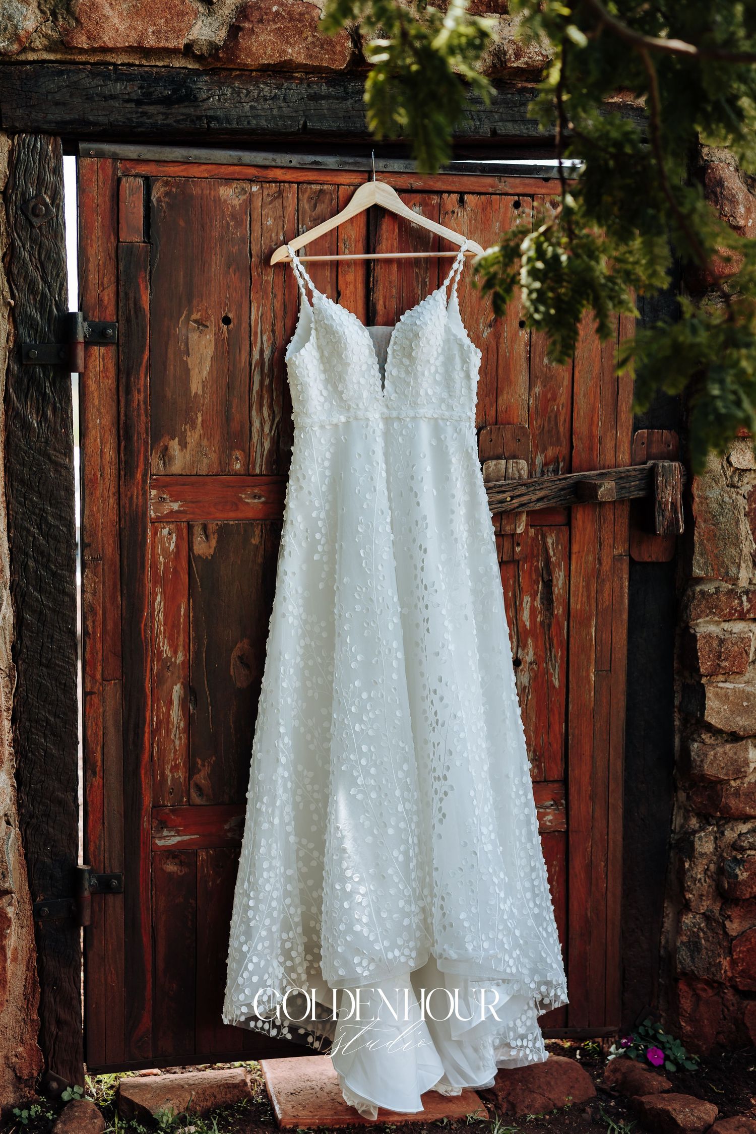 A delicate white lace wedding dress with spaghetti straps hangs on a wooden door.
