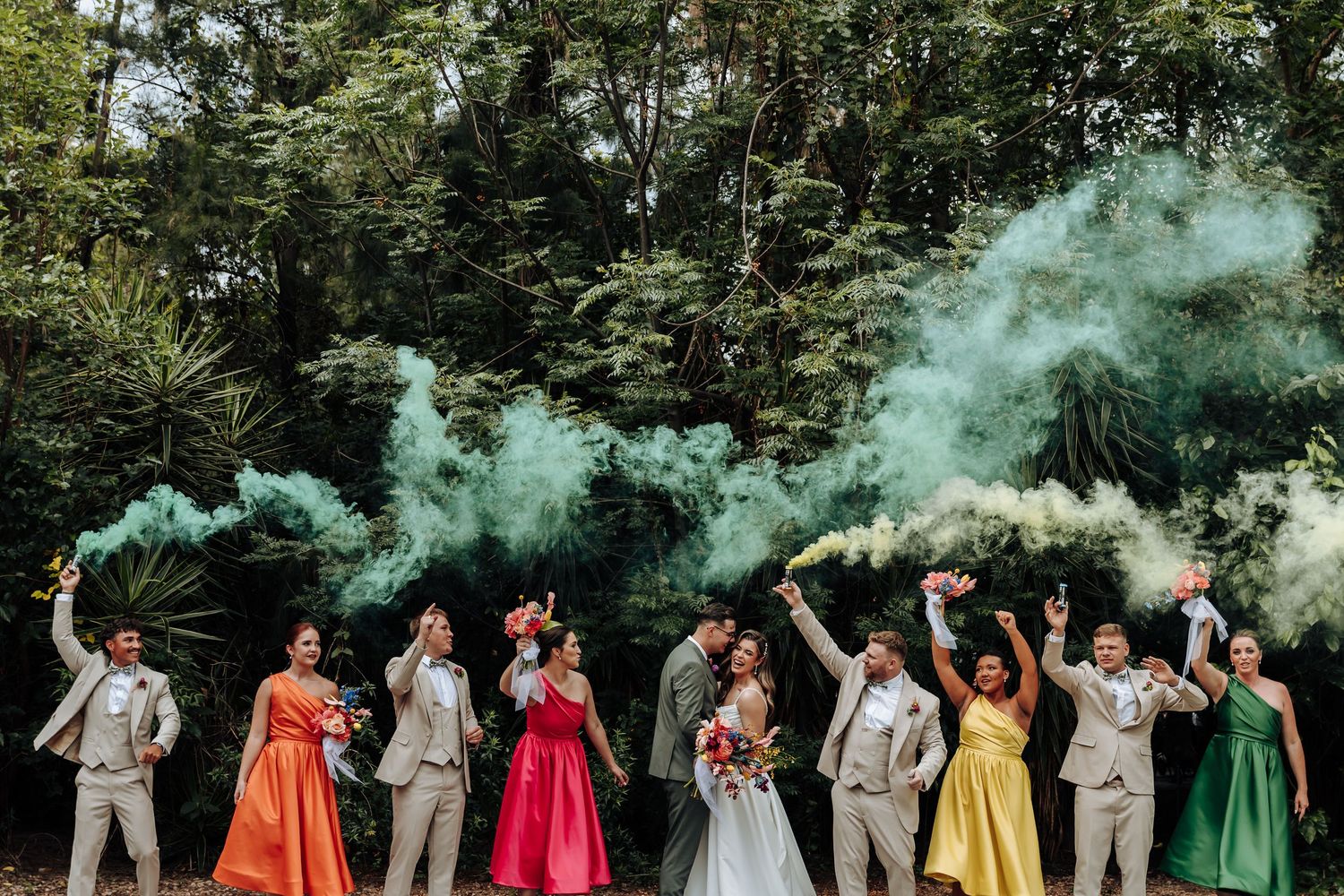 Wedding party members release colorful smoke bombs in a rainbow sequence while standing outdoors among trees.