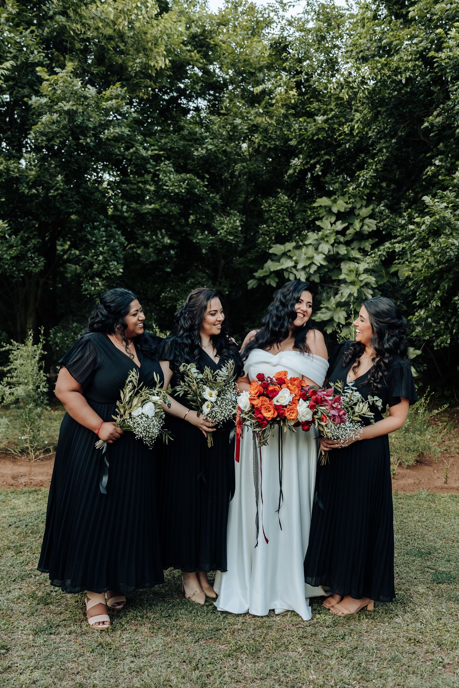 Bridal party in black dresses and white gown sitting together on grass with colorful bouquets.