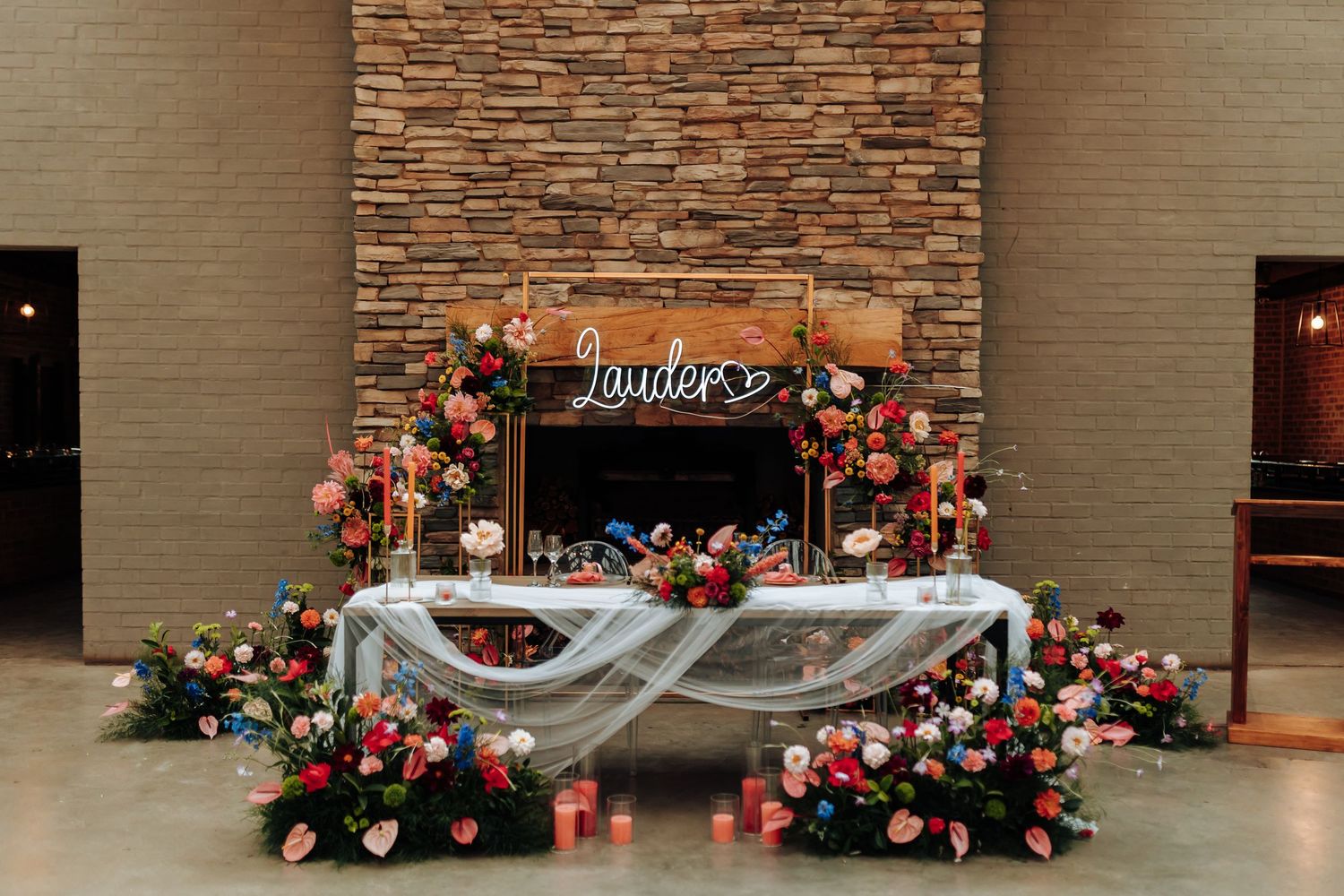 A rustic wedding dessert table with wooden backdrop and pink floral arrangements against a stone brick wall.