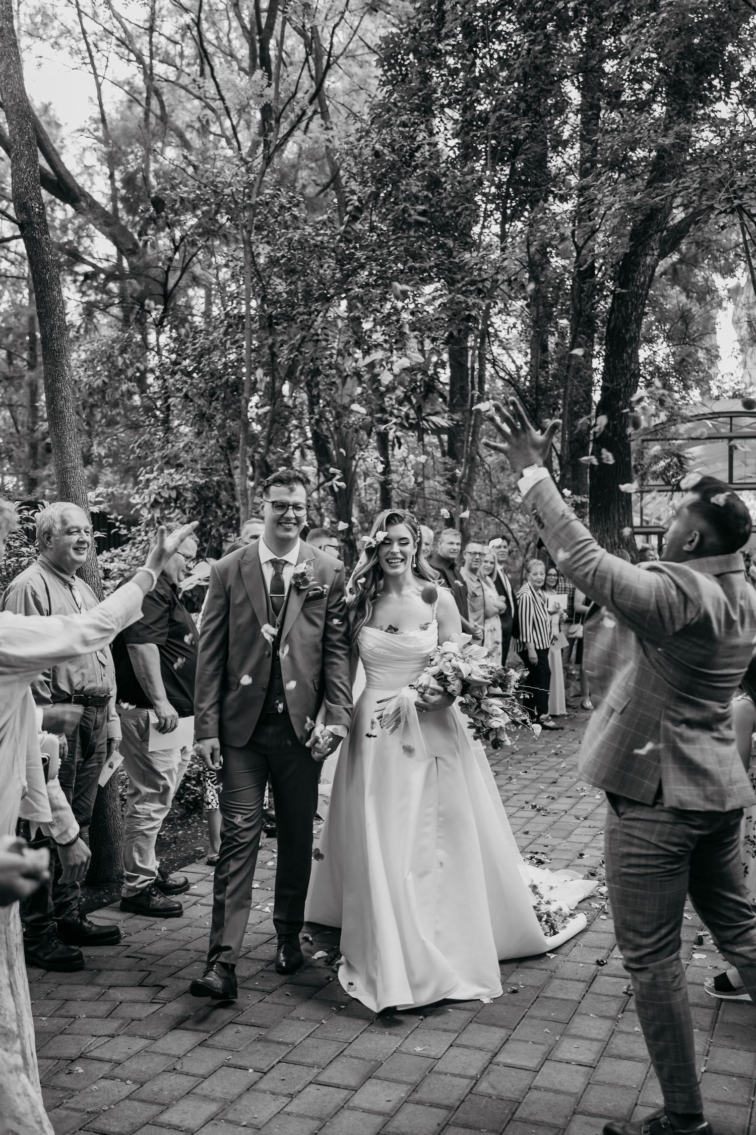 Wedding party celebrates outdoors on a brick pathway surrounded by trees.