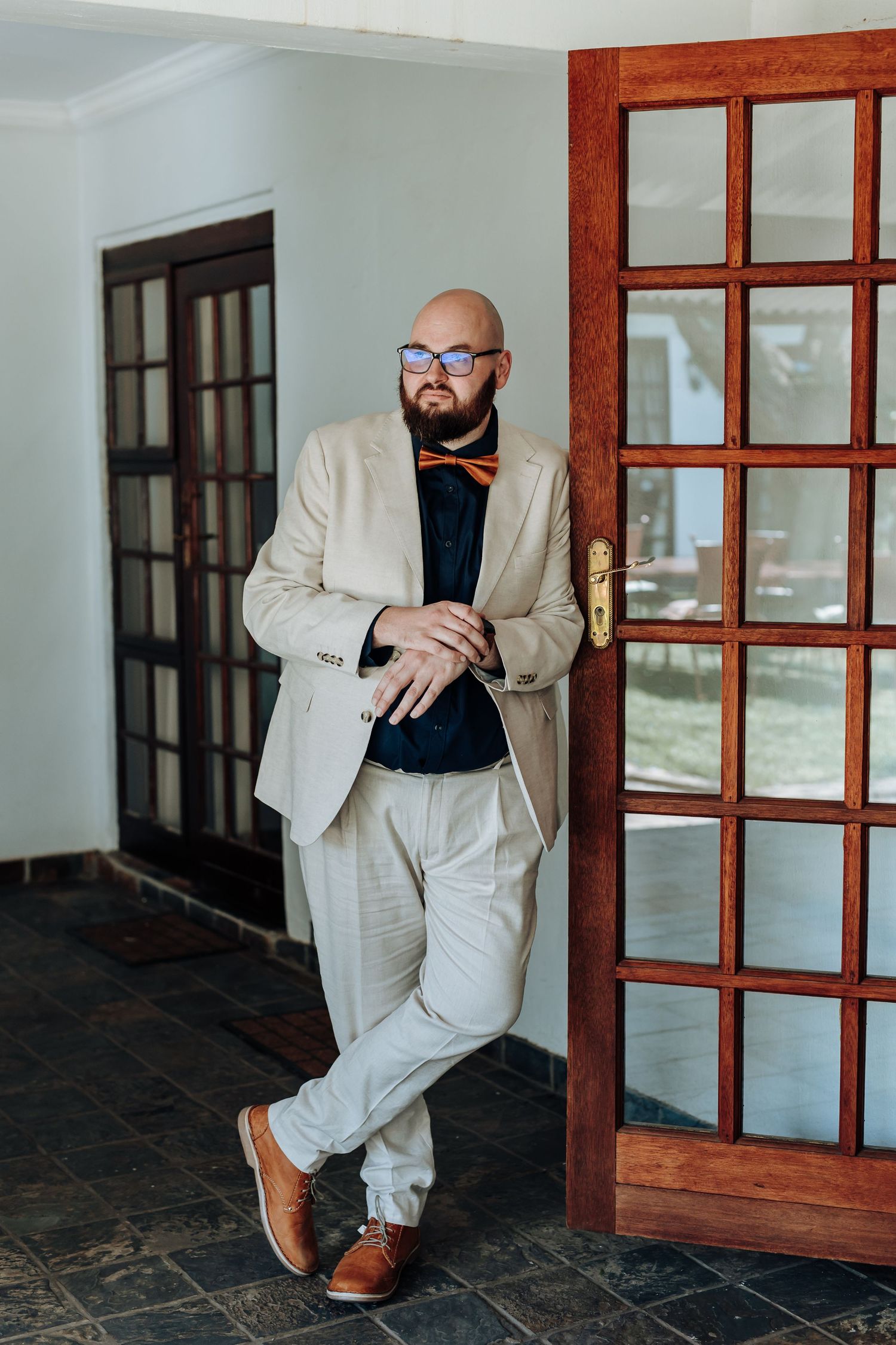 A person in a beige suit and brown shoes sits on a dark tiled floor next to wooden doors in an indoor hallway.