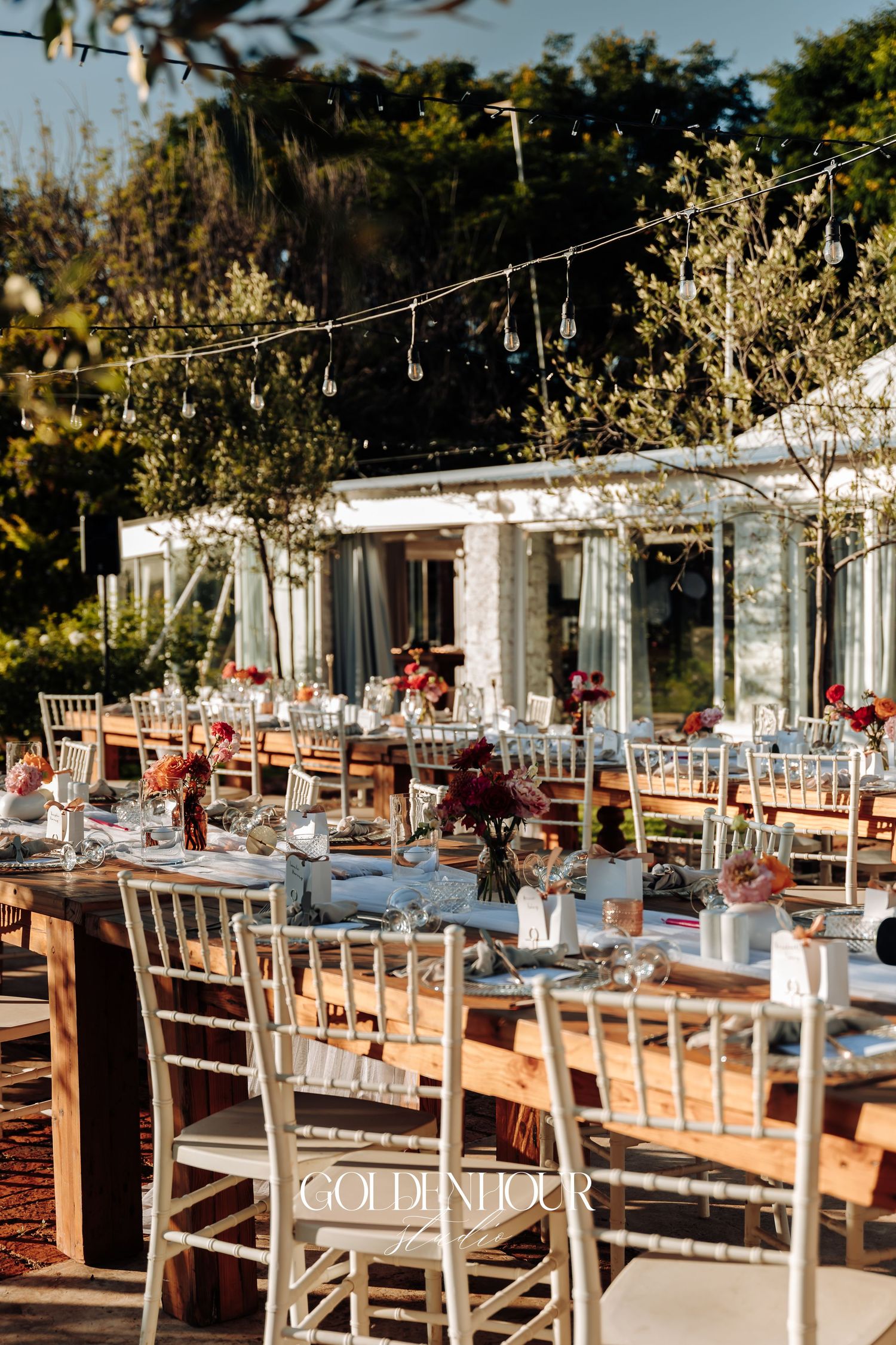 Elegant outdoor wedding reception setup with white tables, wooden chairs, and rustic decor under natural sunlight.