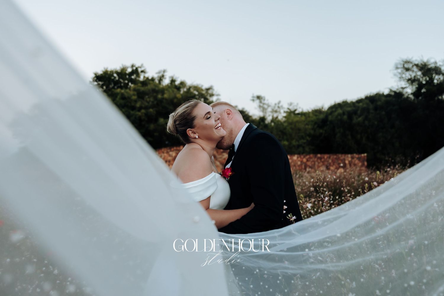 A couple shares a romantic kiss outdoors while a flowing white veil catches the breeze.
