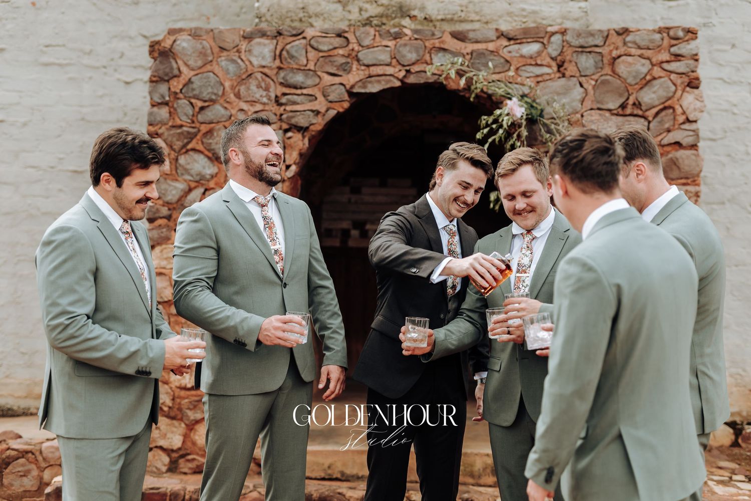 Groomsmen in gray suits share drinks and laughs during outdoor wedding celebration.