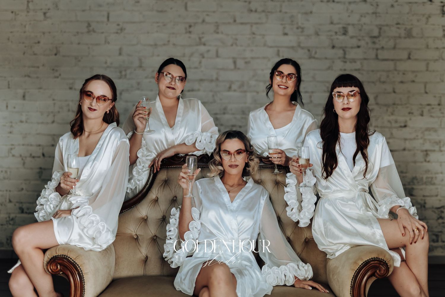 Group photo on beige tufted sofa wearing matching white silk robes against brick wall.