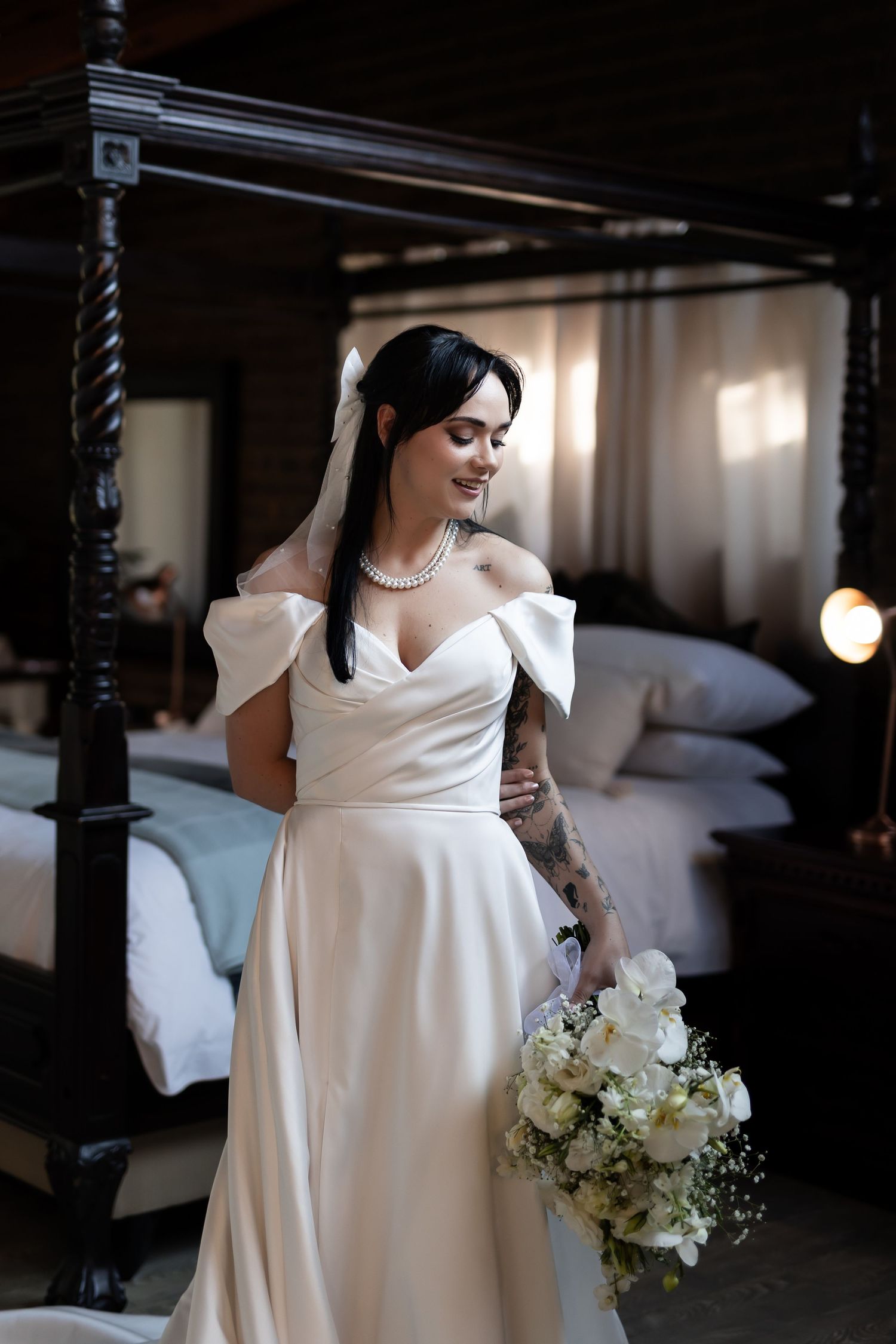 Elegant off-shoulder white wedding dress with floral bouquet in moody indoor setting near classic four-poster bed.