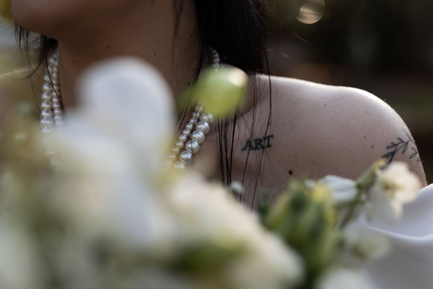 Pearl necklace and shoulder tattoo visible in soft focus with blurred white flowers in foreground.
