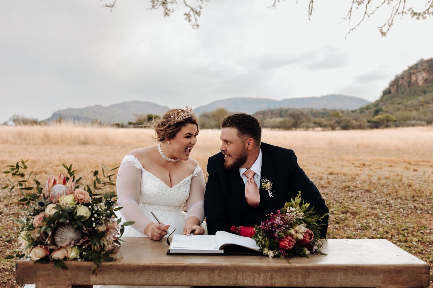 Couple signing paperwork at rustic wooden table in desert landscape with mountain backdrop and floral arrangements.