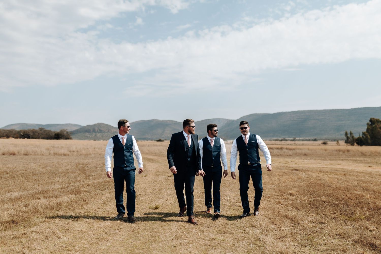 Groomsmen in dark suits stand together against a golden field and mountain landscape on a sunny day.