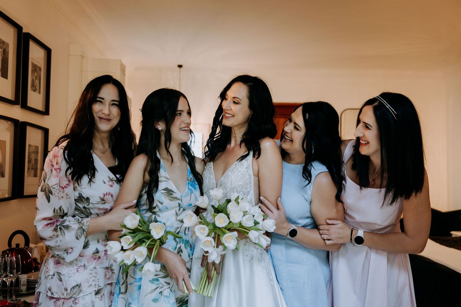 Bride holding white tulip bouquet laughing with four bridesmaids in blue and floral dresses during wedding preparations.