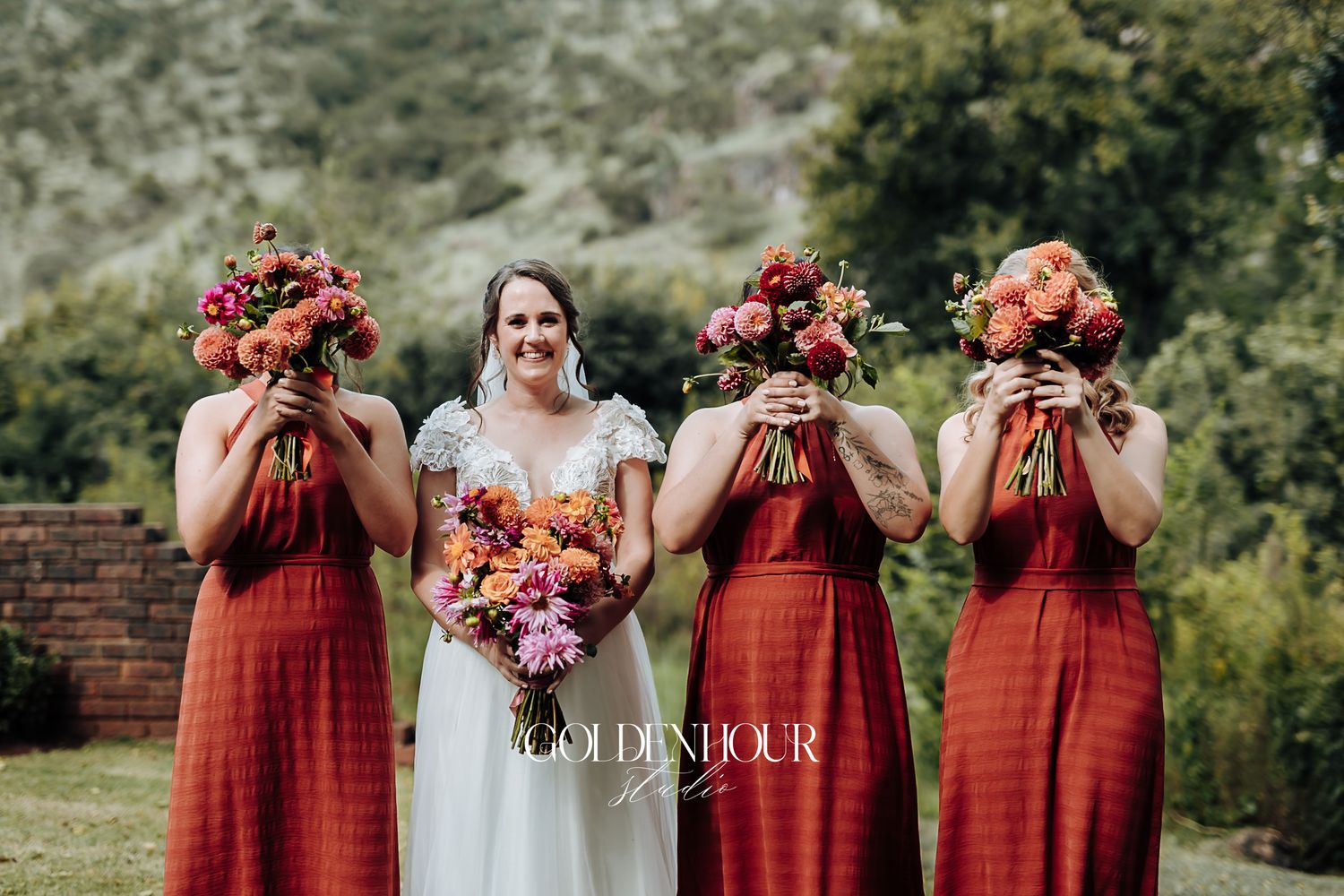 Bride and three bridesmaids in rust dresses hiding faces behind vibrant wedding bouquets outdoors.