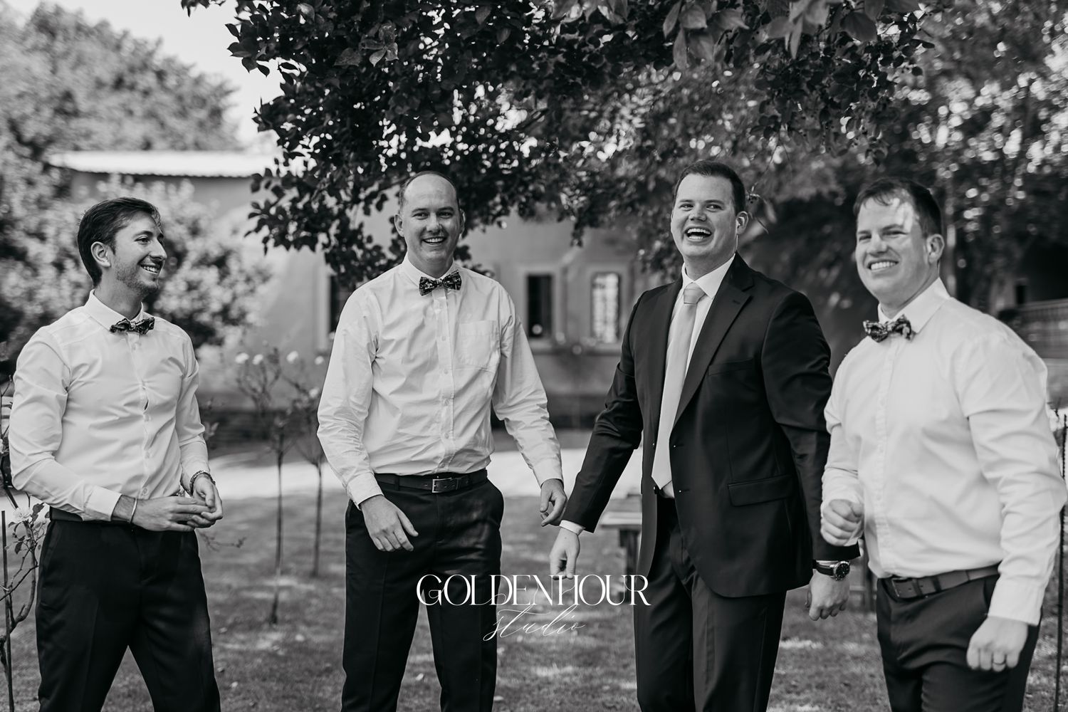 Black and white photo of four groomsmen laughing together outdoors at a wedding celebration.